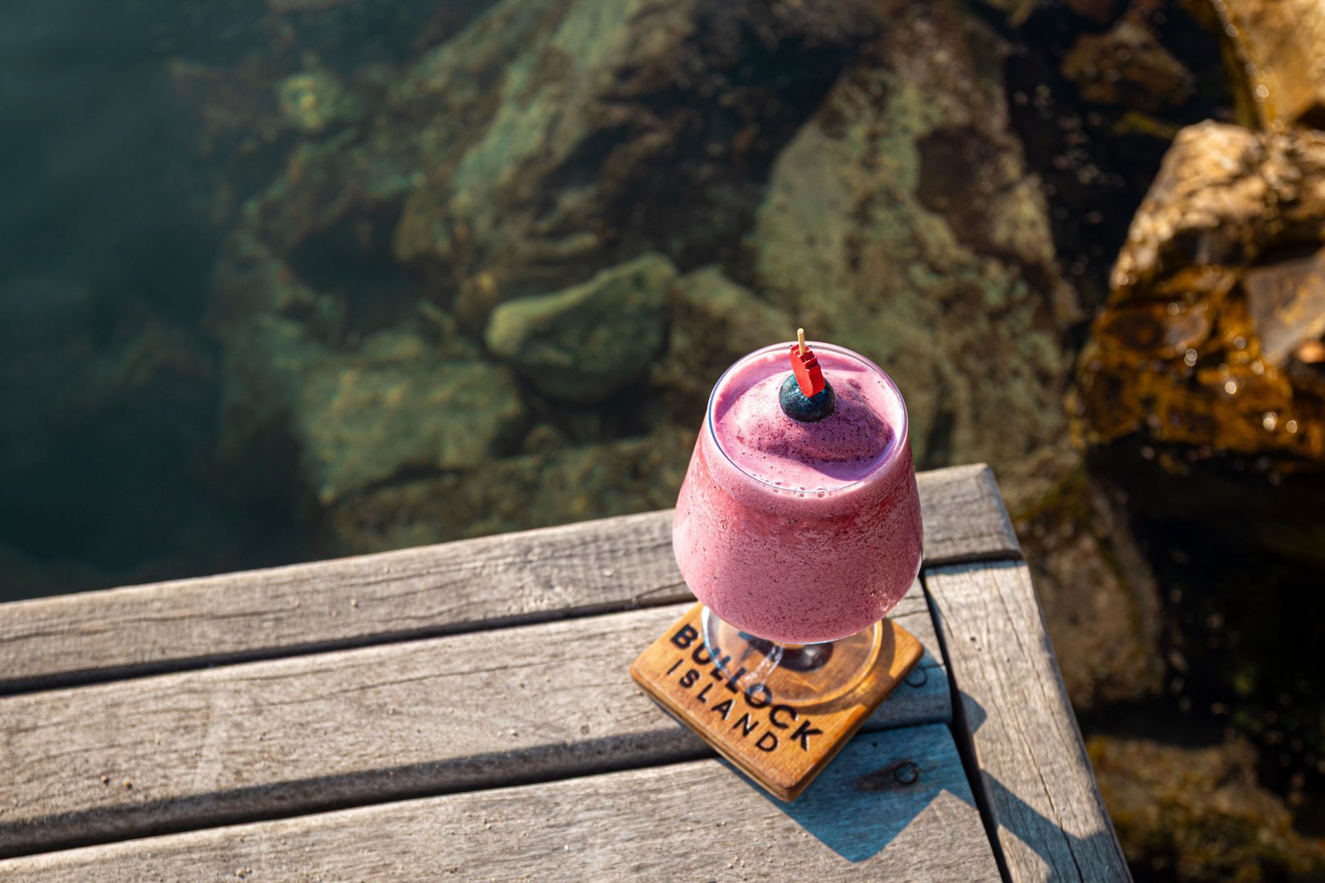 Pink layered dessert with a cherry on top on a wooden deck beside clear water
