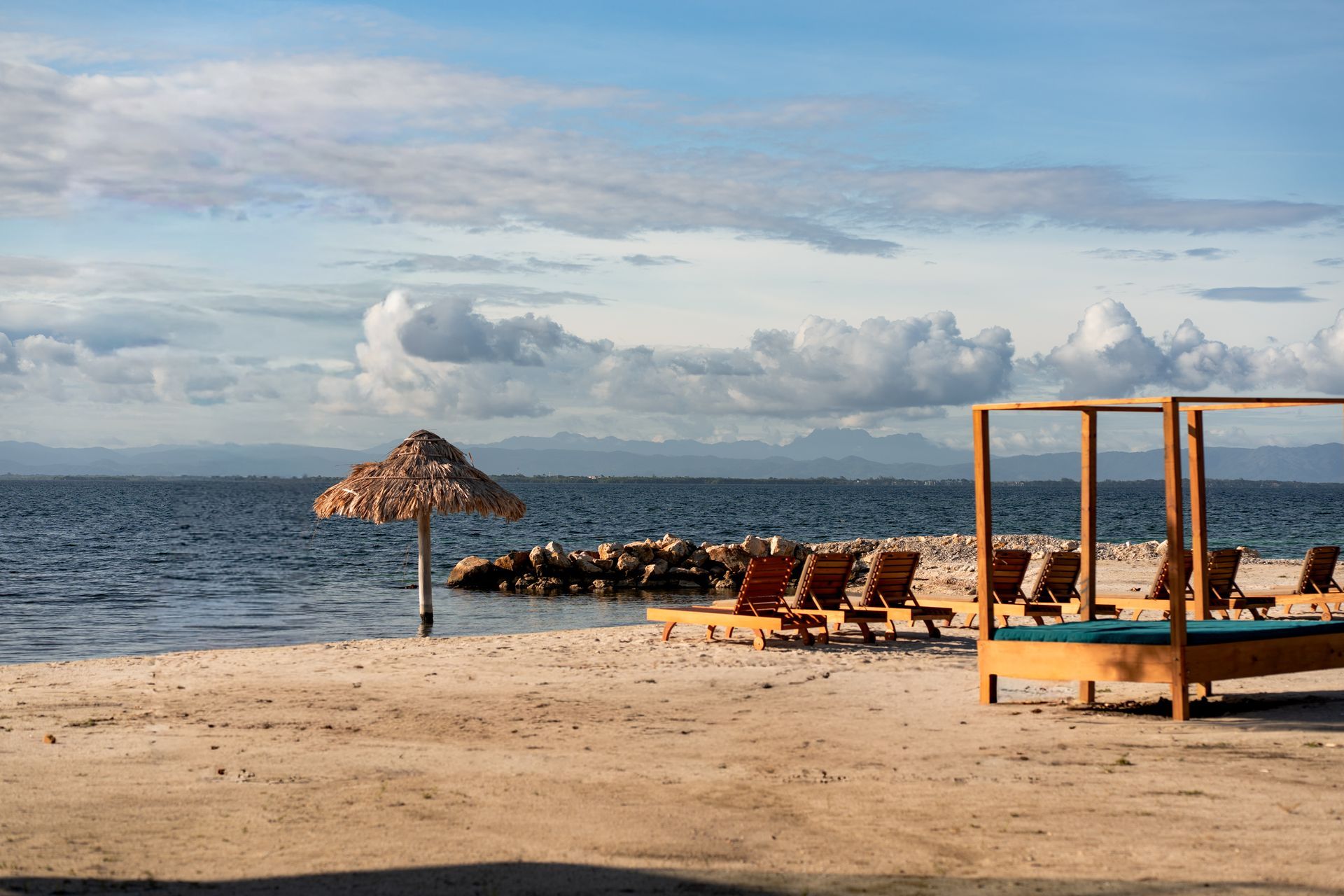 Sandy beach with lounge chairs, thatched umbrella, and wooden cabanas beside a calm blue sea