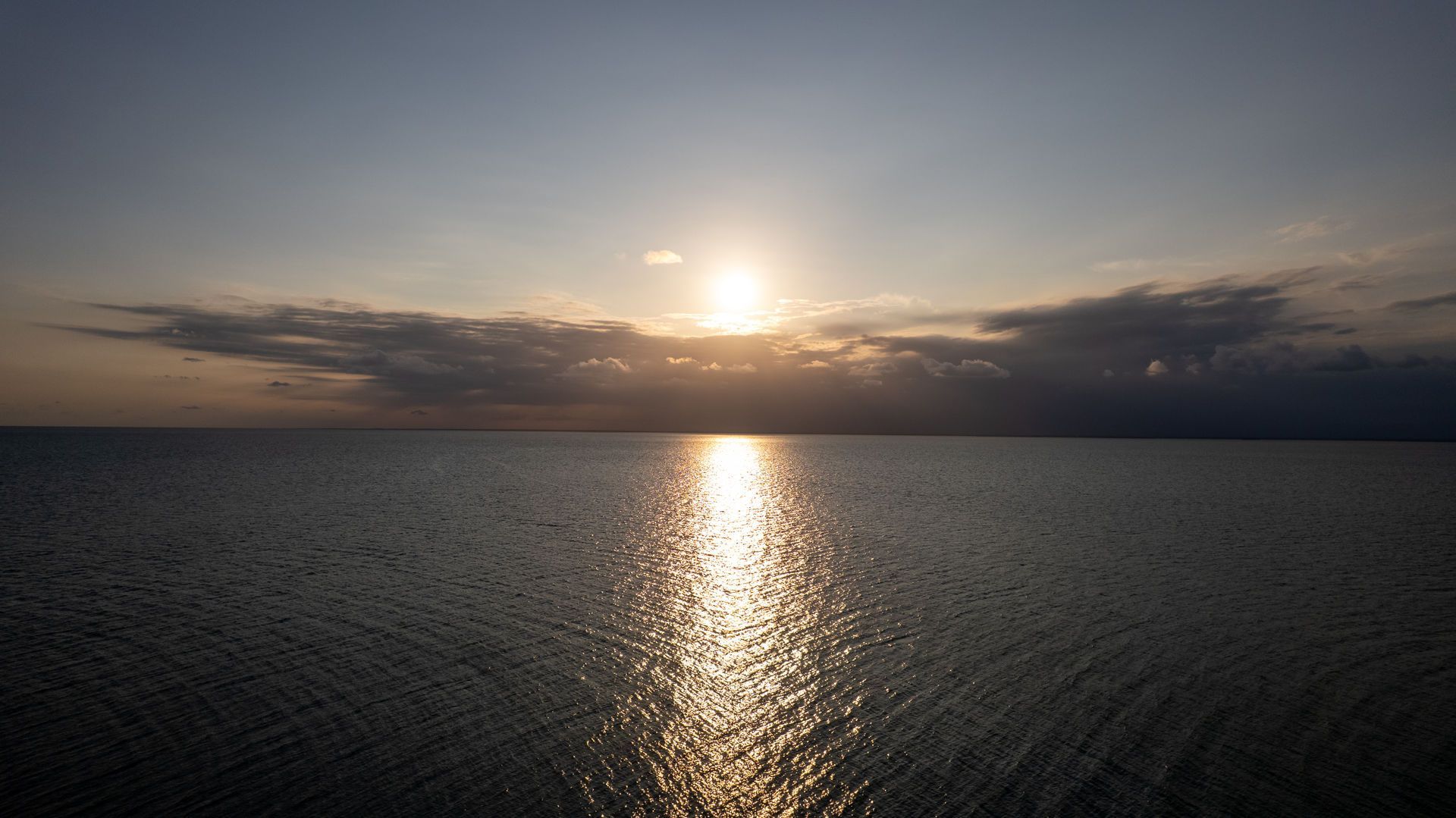 Sunset over calm ocean, with a bright reflection on the water beneath a cloudy sky.