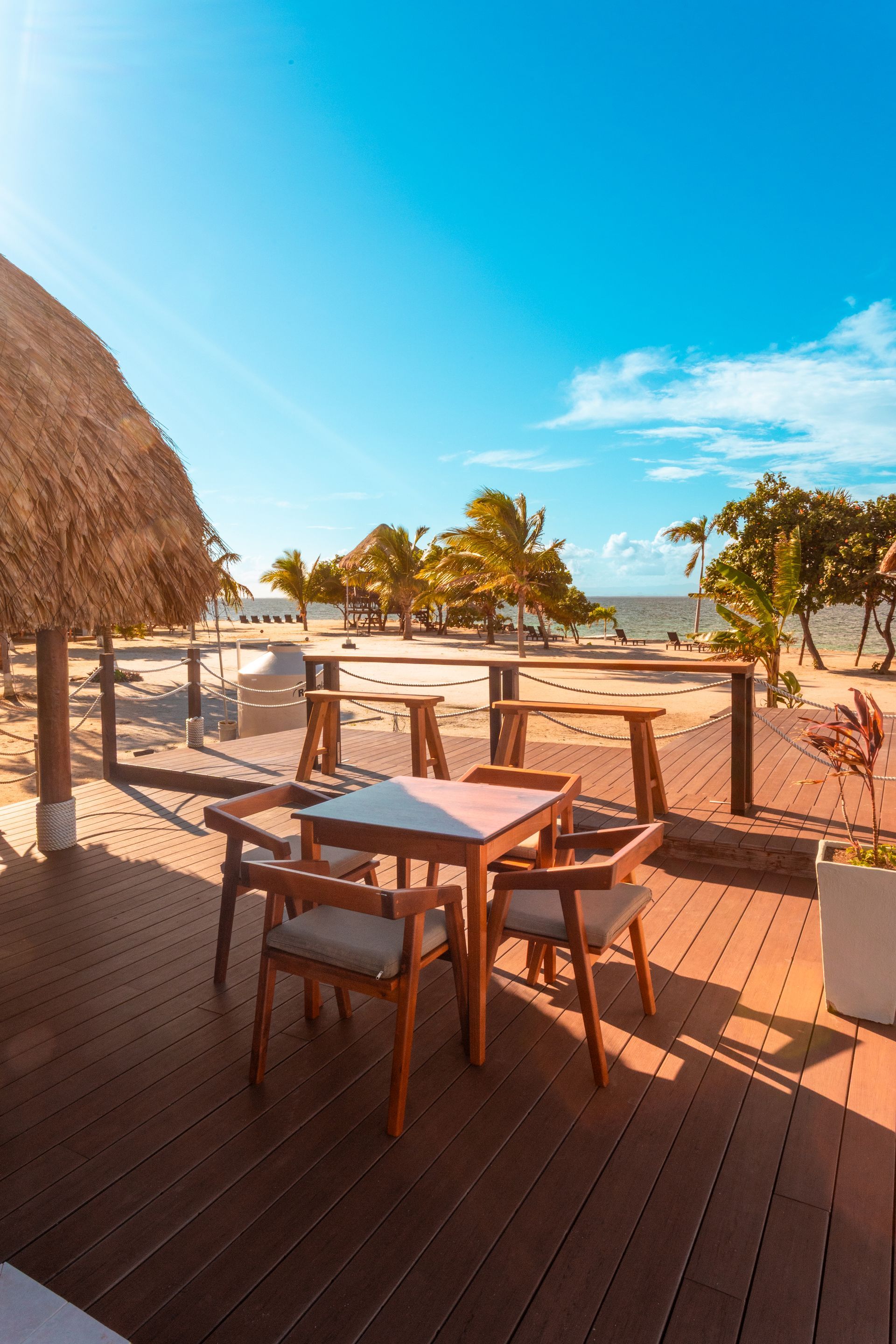 Beachfront patio with wooden tables and chairs under a thatched roof, overlooking sand and palm trees at sunset