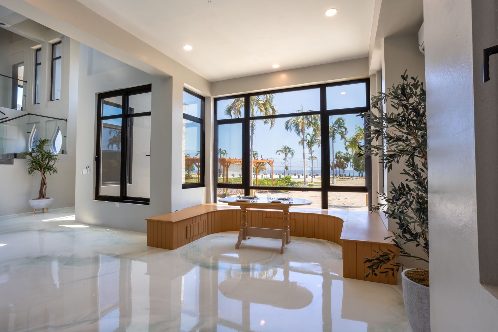 Bright modern living room with glossy white floors, beige furniture, and large windows overlooking trees and water.