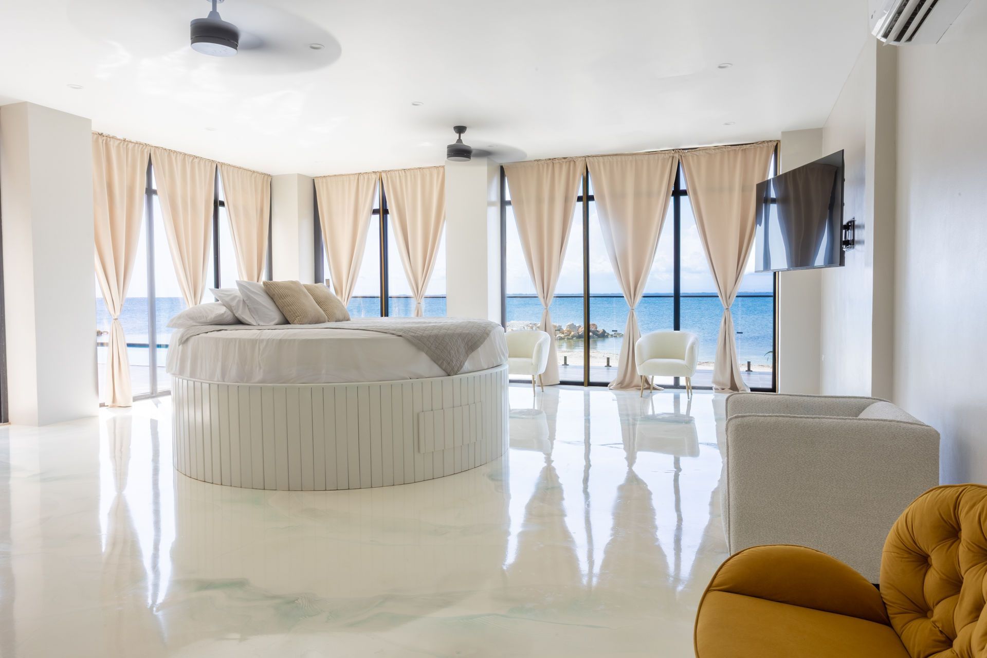 Bright bedroom with a round bed, white floors, beige curtains, and ocean views through large windows