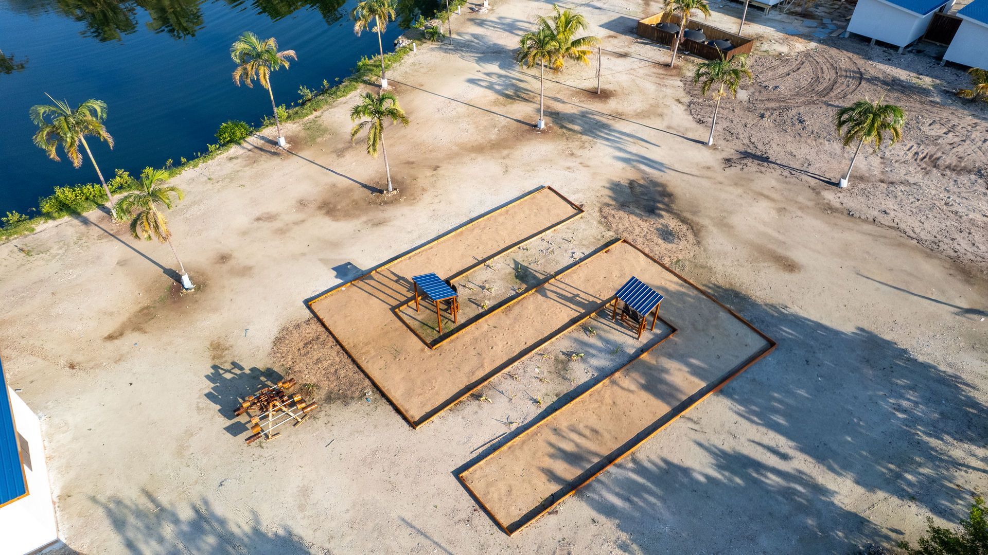 Aerial view of two empty picnic tables on sandy ground near blue water and scattered palm trees