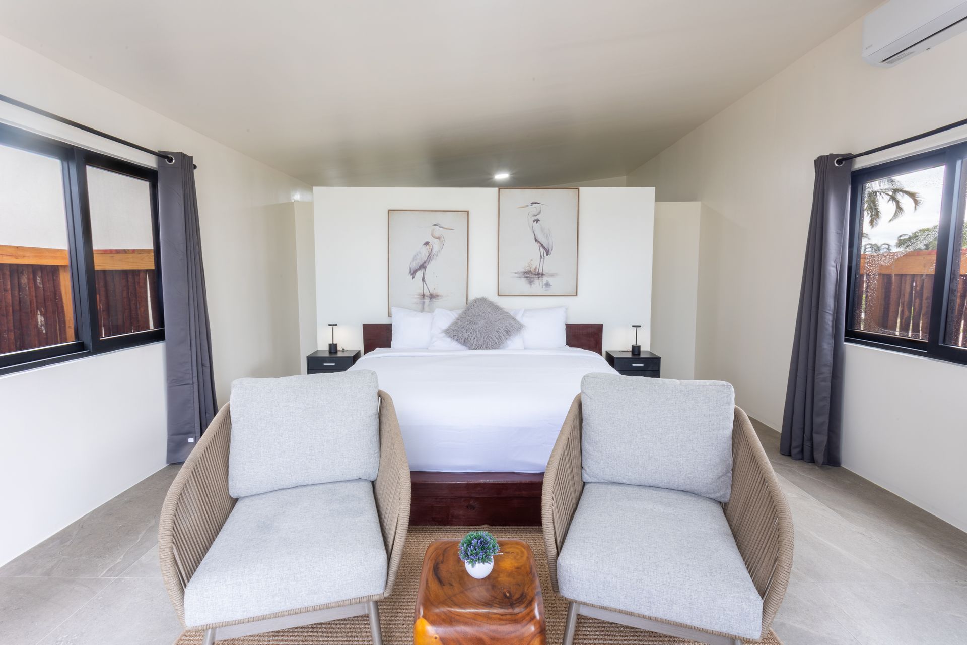Modern bedroom with white bed, two wicker chairs, and large windows with gray curtains