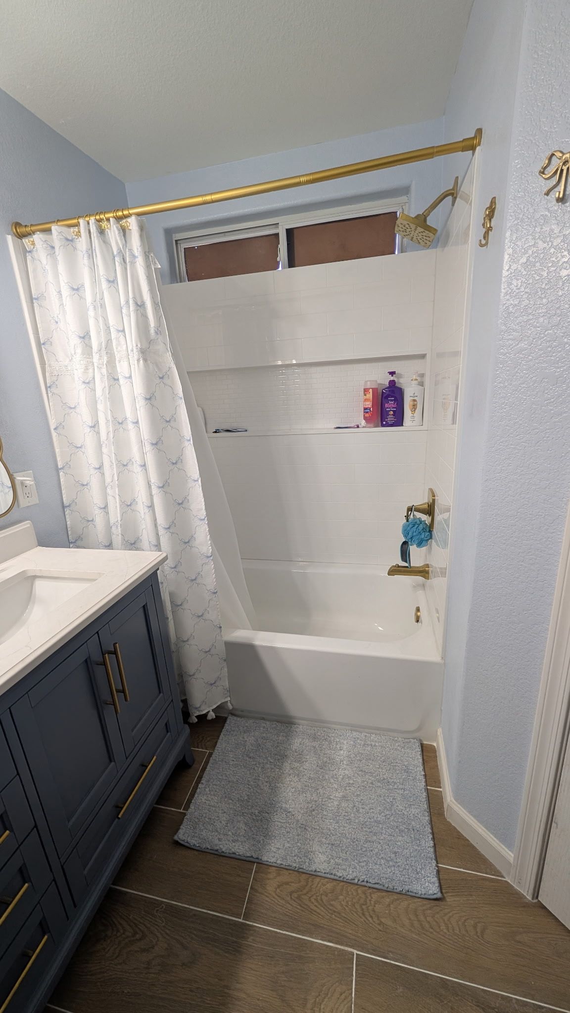 White shower/tub alcove with curtain, gold rod, small shelf, bath mat, and gray cabinet in a bathroom