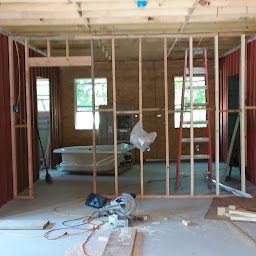 A wooden frame wall under construction in a room, featuring a bathtub, a red stepladder, and a circular saw on the floor.