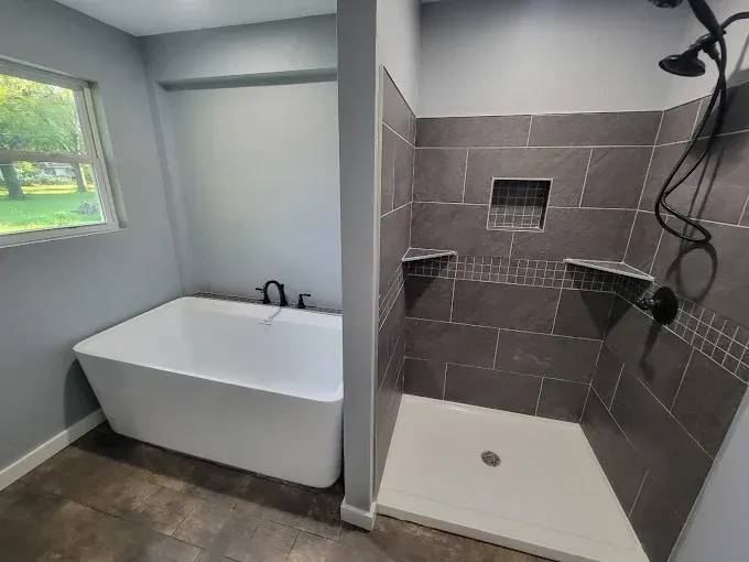 Bathroom with a white soaking tub, walk-in shower, gray tile, and a window