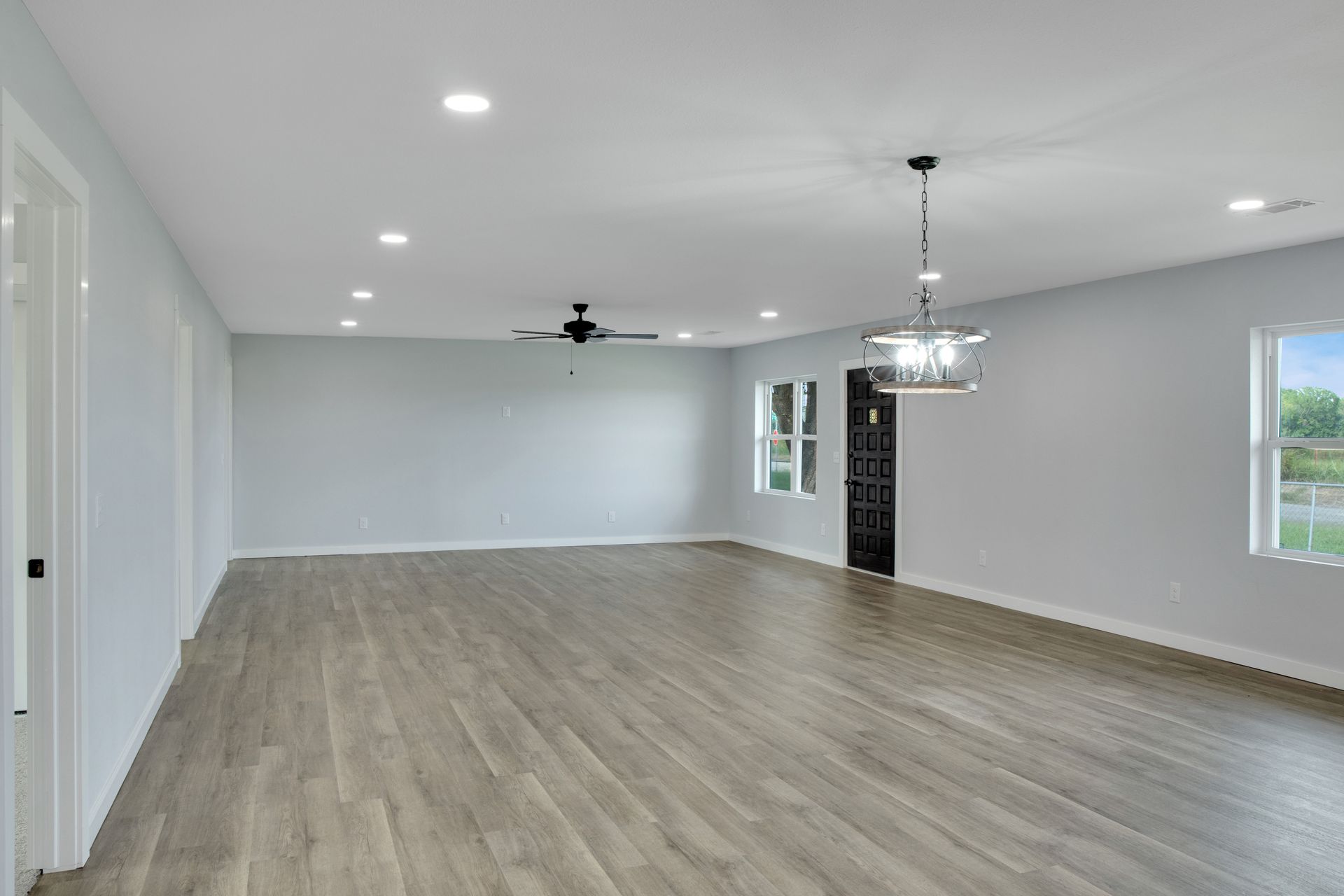 An empty, spacious room with light gray walls, light wood-look flooring, recessed lighting, a ceiling fan, and chandelier.