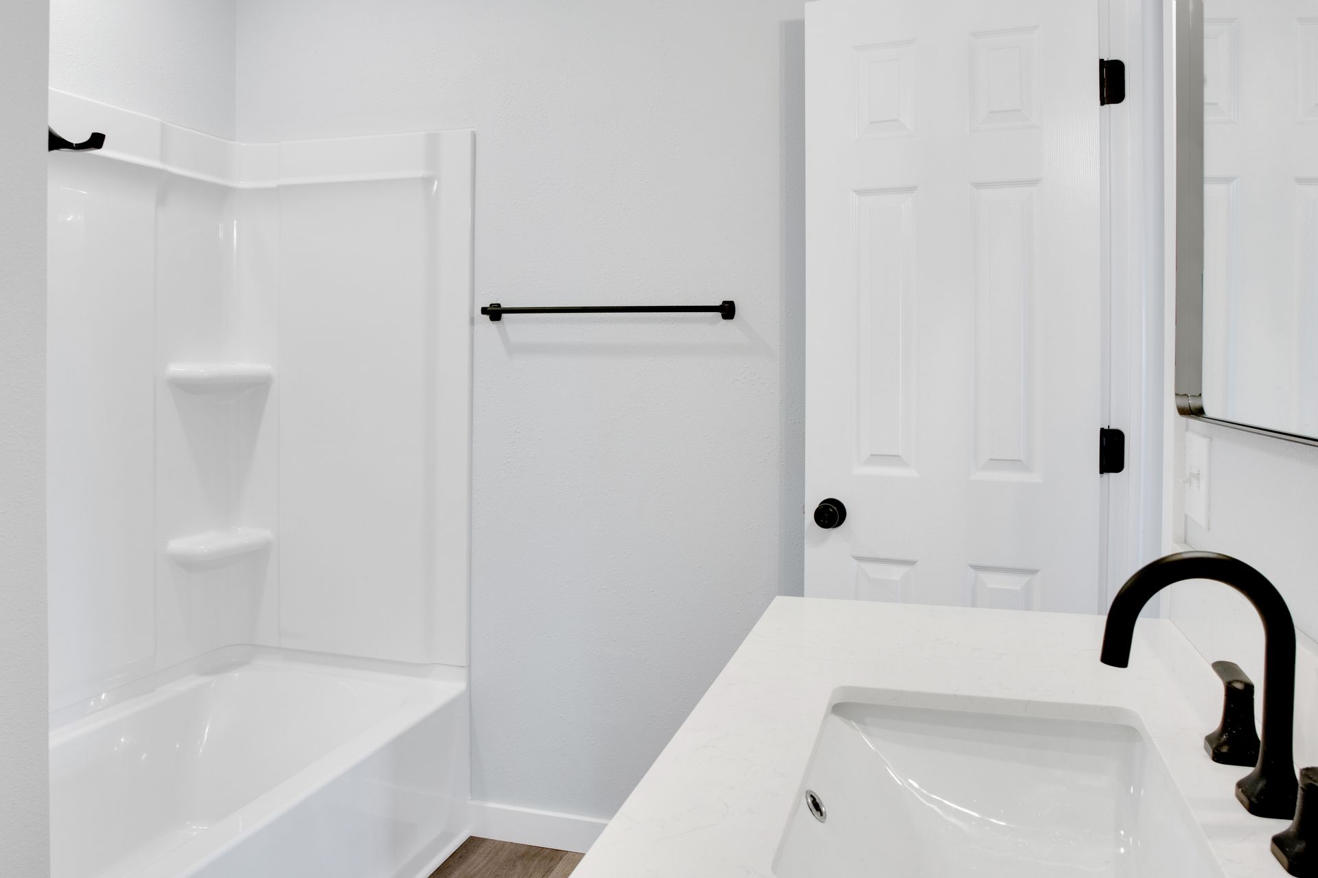 A modern bathroom featuring a white tub-shower combo, a light gray wall with a black towel bar, and a white vanity.