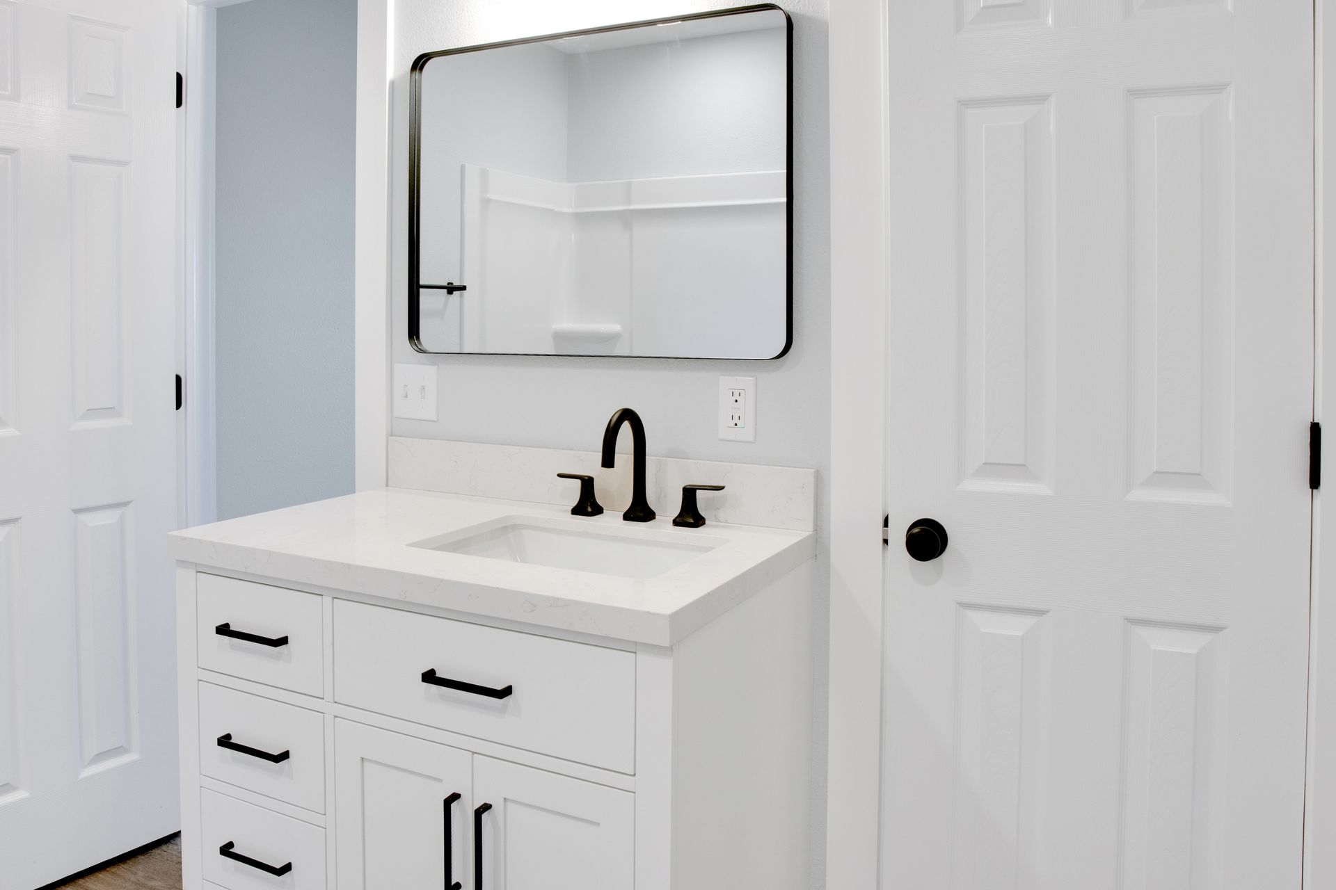 A white bathroom vanity with a black faucet, rectangular mirror, and black hardware against white walls.