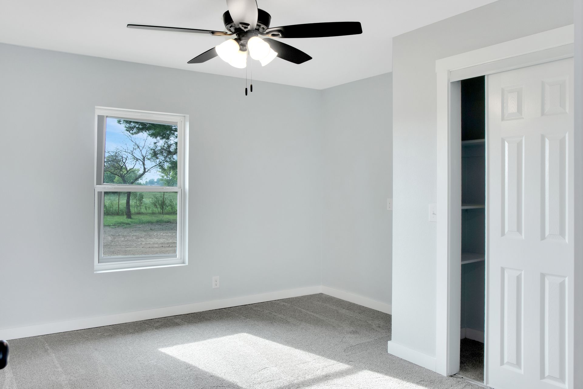 An empty bedroom with light gray walls, gray carpet, a ceiling fan, and a partially open white closet door.