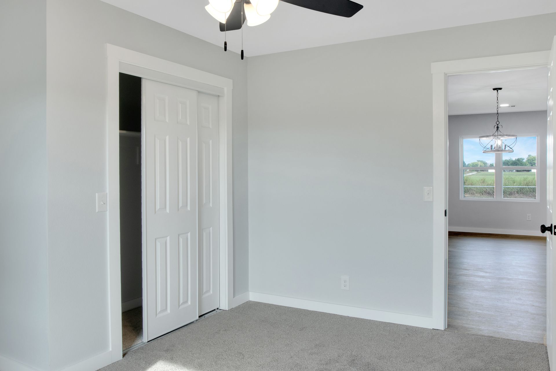 A light gray bedroom with carpet, a white closet with sliding doors, and a doorway looking into an adjacent room.