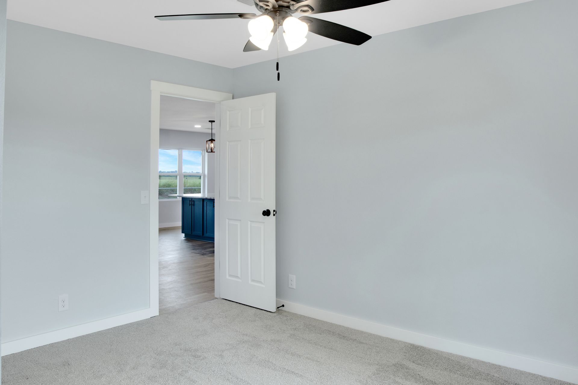 An empty bedroom with light gray walls, gray carpet, a white door, and a ceiling fan, with a doorway leading to a kitchen.