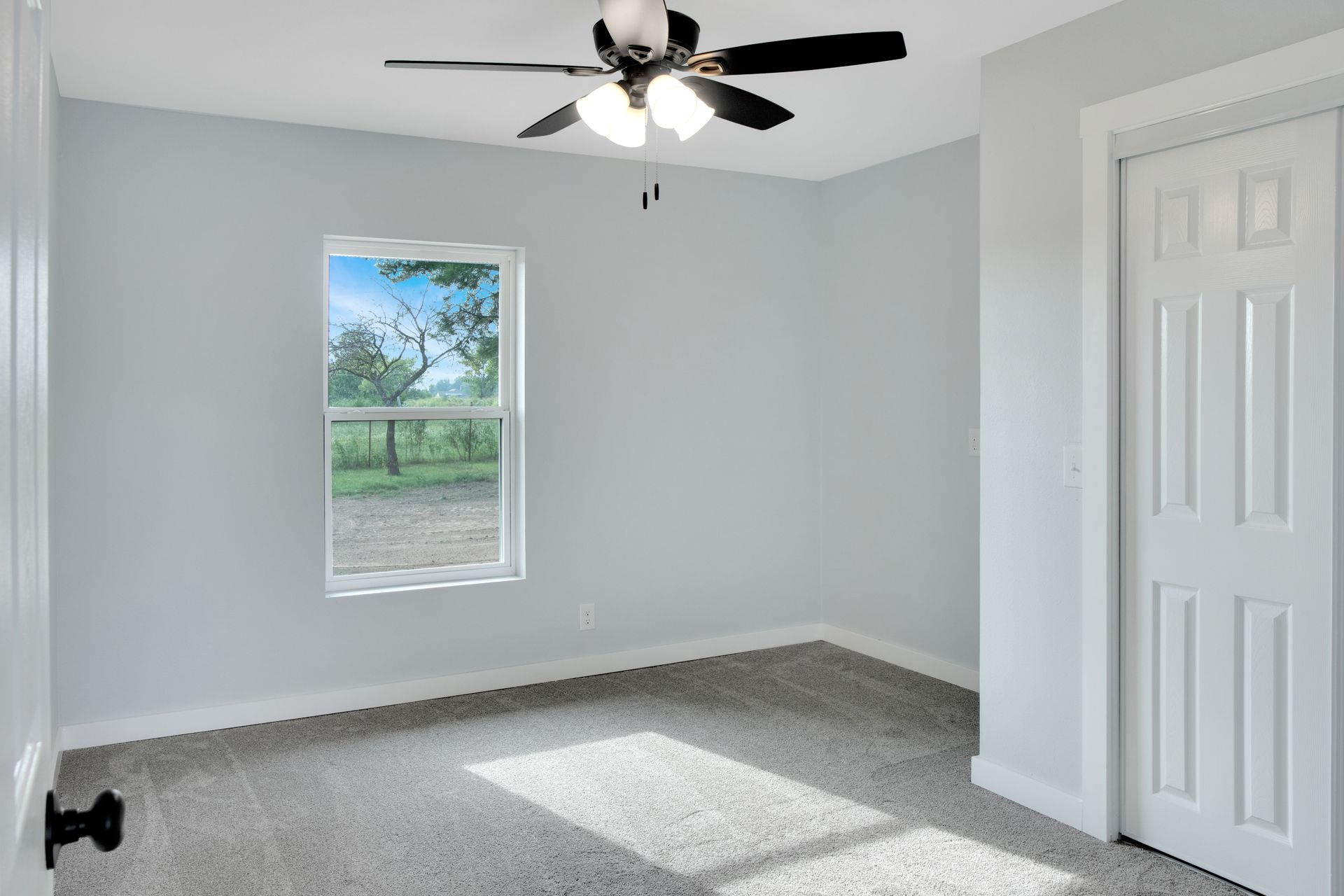 Empty bedroom with light grey walls, white trim, grey carpet, a ceiling fan, and a single window showing green outdoors.