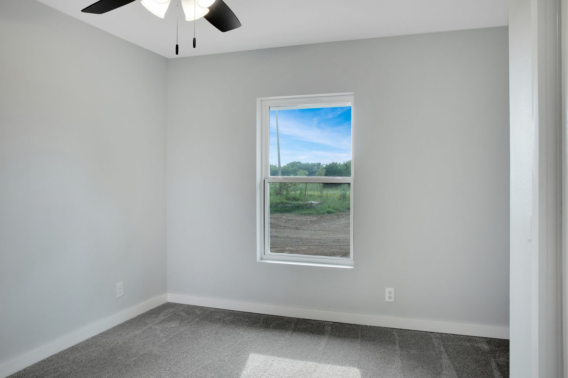 A view of an empty bedroom with light gray walls, gray carpet, a white-framed window, and a black ceiling fan.