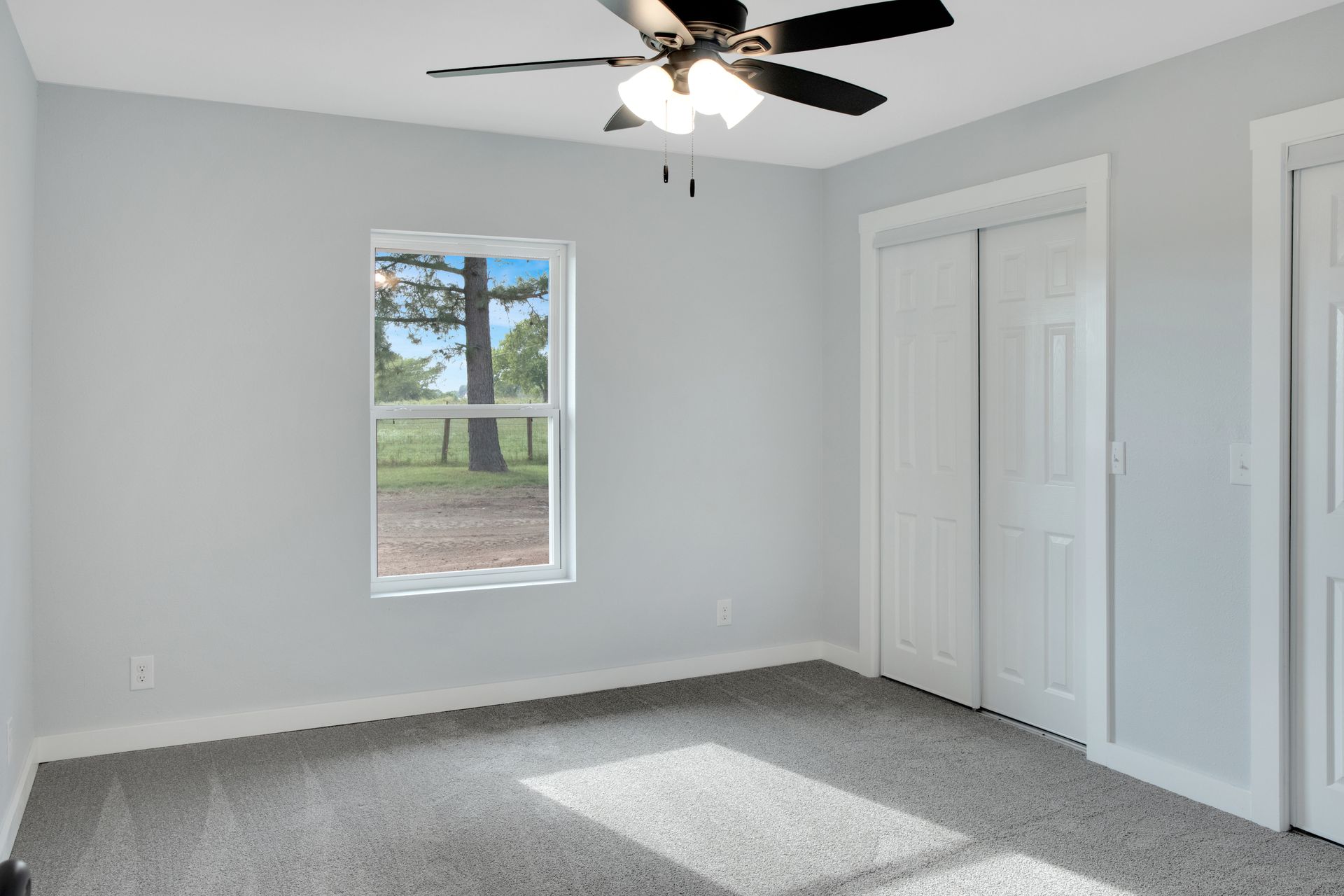 A bright, empty bedroom with light gray walls, gray carpet, a single window, a white ceiling fan, and a white closet.