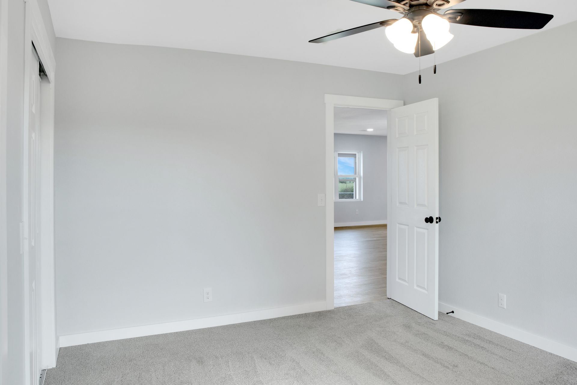 A room with light grey walls, grey carpet, a ceiling fan, and an open white door leading into a hallway with a window.