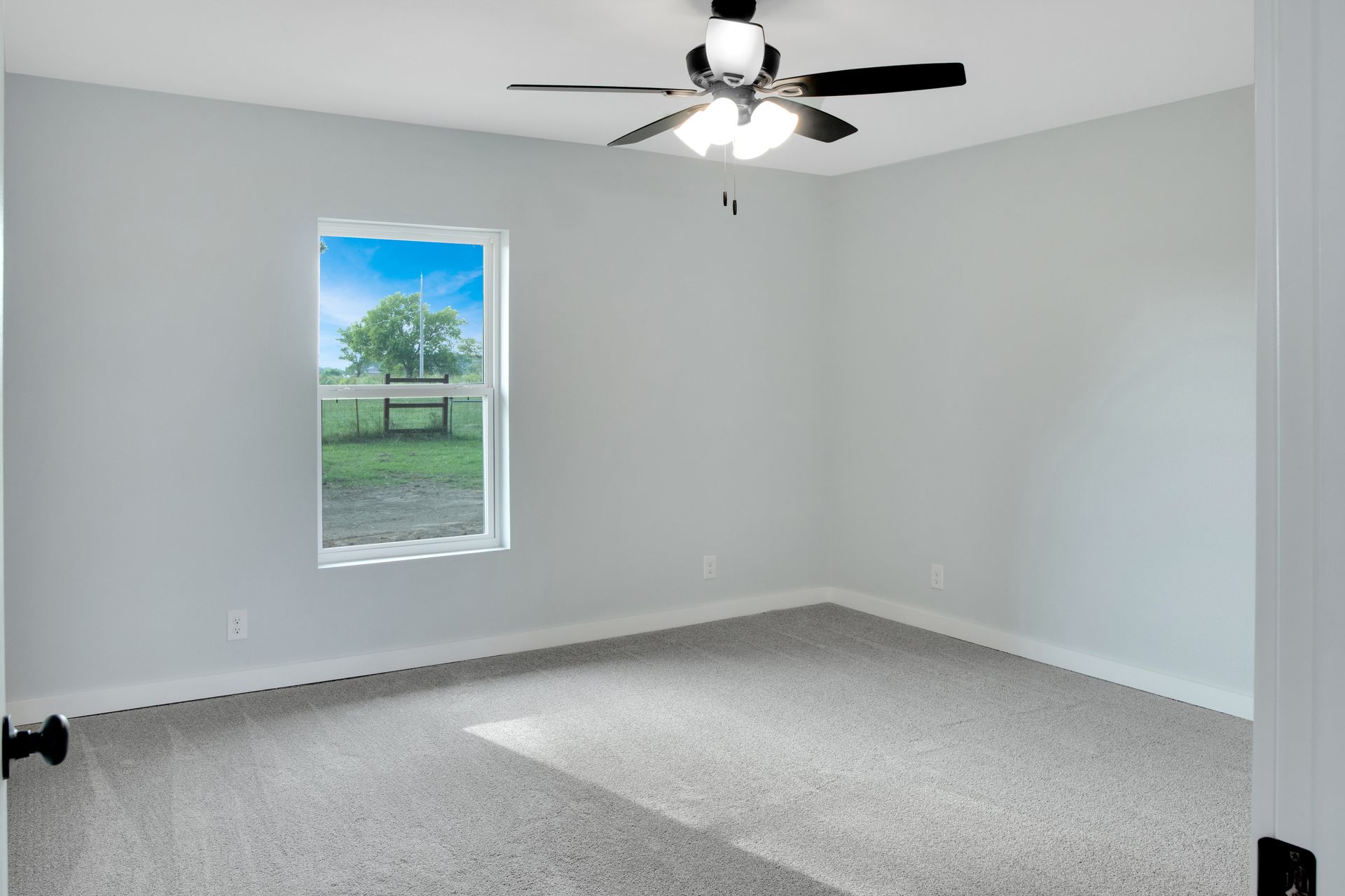An empty bedroom with light gray walls, plush gray carpet, a single window, and a white ceiling fan with light fixtures.