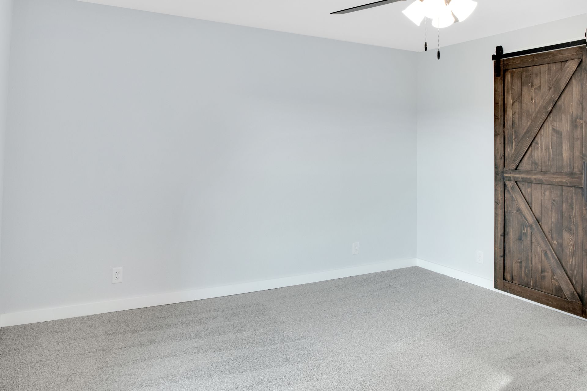 An empty room with light gray walls, gray carpet, a ceiling fan, and a dark wood sliding barn door on the right.