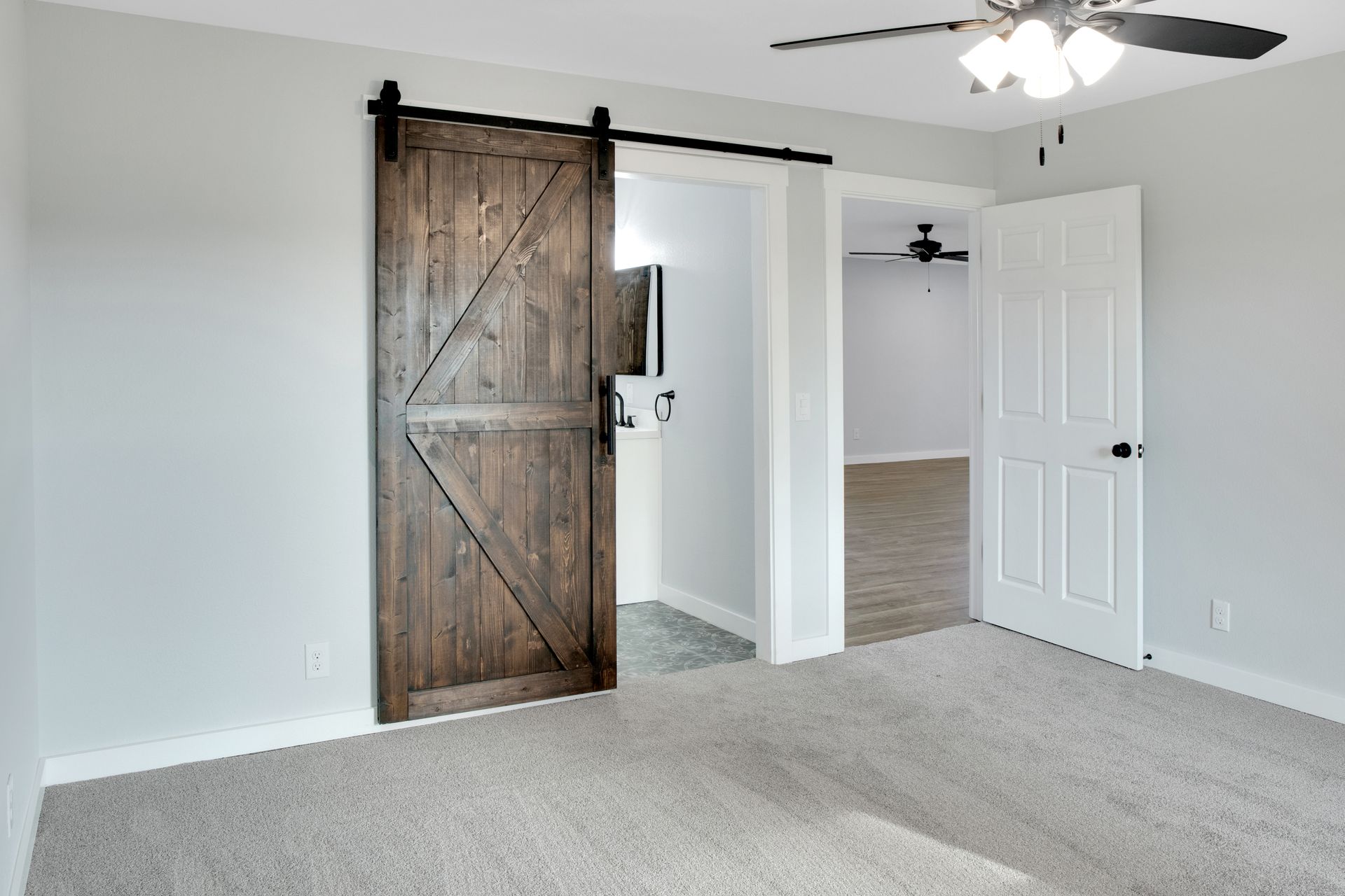 A room with light grey walls, grey carpet, a wooden barn door leading to a bathroom, and a white interior door.