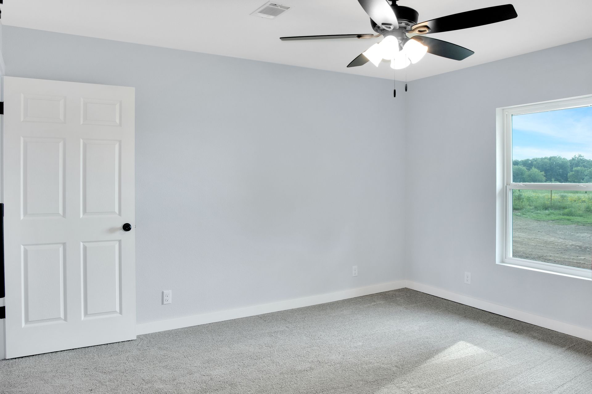 An empty bedroom with light gray walls, gray carpet, a white door, a ceiling fan, and a window overlooking a grassy area.