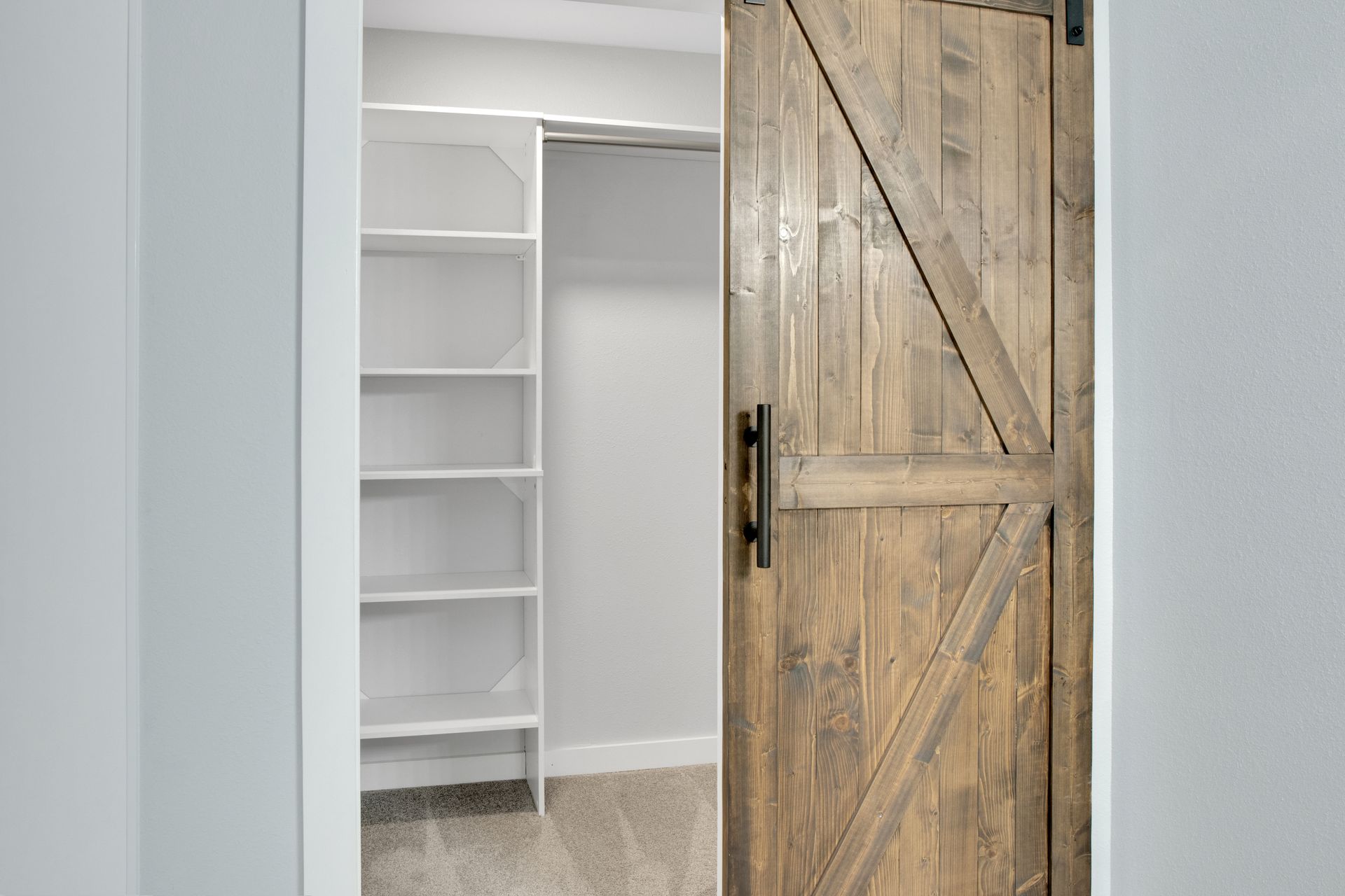 A sliding wooden barn door partially open, revealing a walk-in closet with white shelving and beige carpeting.