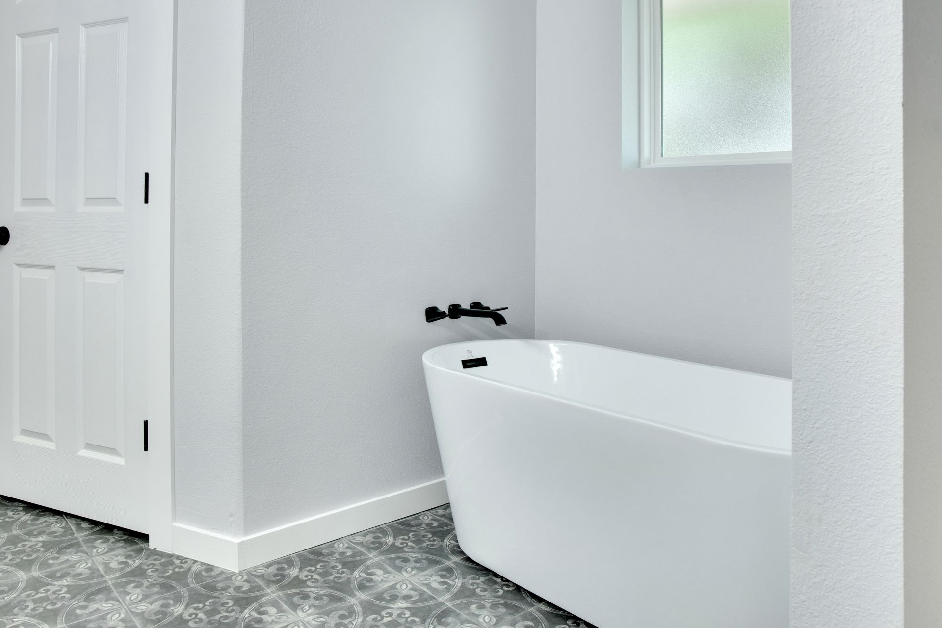A white freestanding bathtub with black hardware against a grey wall in a bathroom with patterned tile flooring.