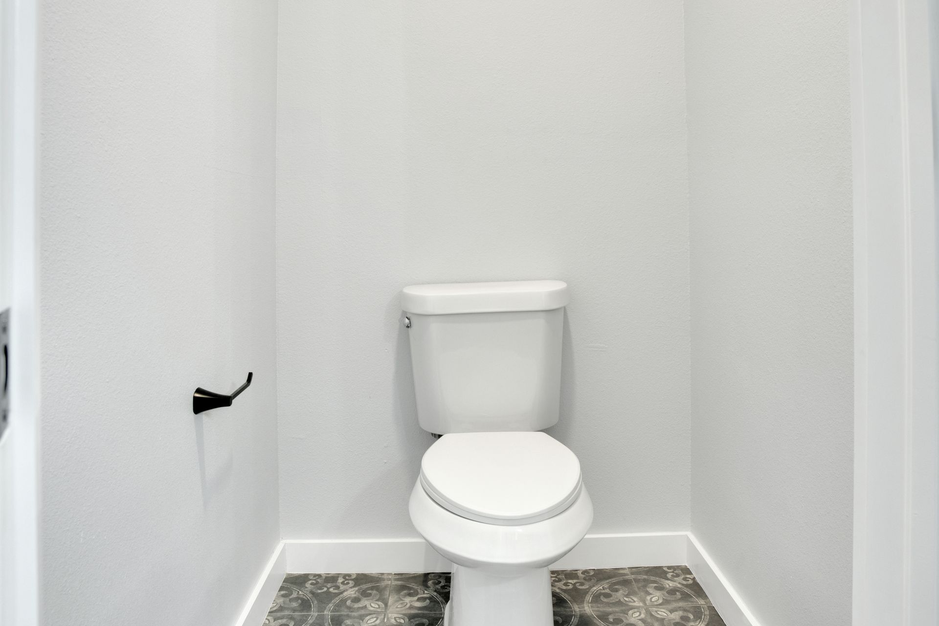 A minimalist water closet with white walls, a white toilet, and patterned grey floor tiles.