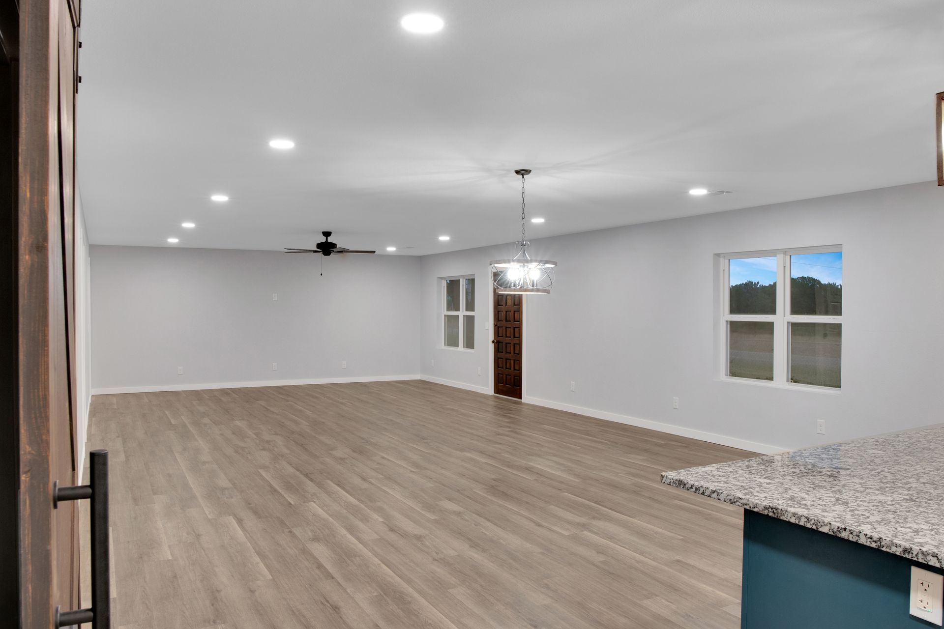 A bright, open-concept living area with light-toned wood floors, grey walls, recessed lighting, and a dark wood door.