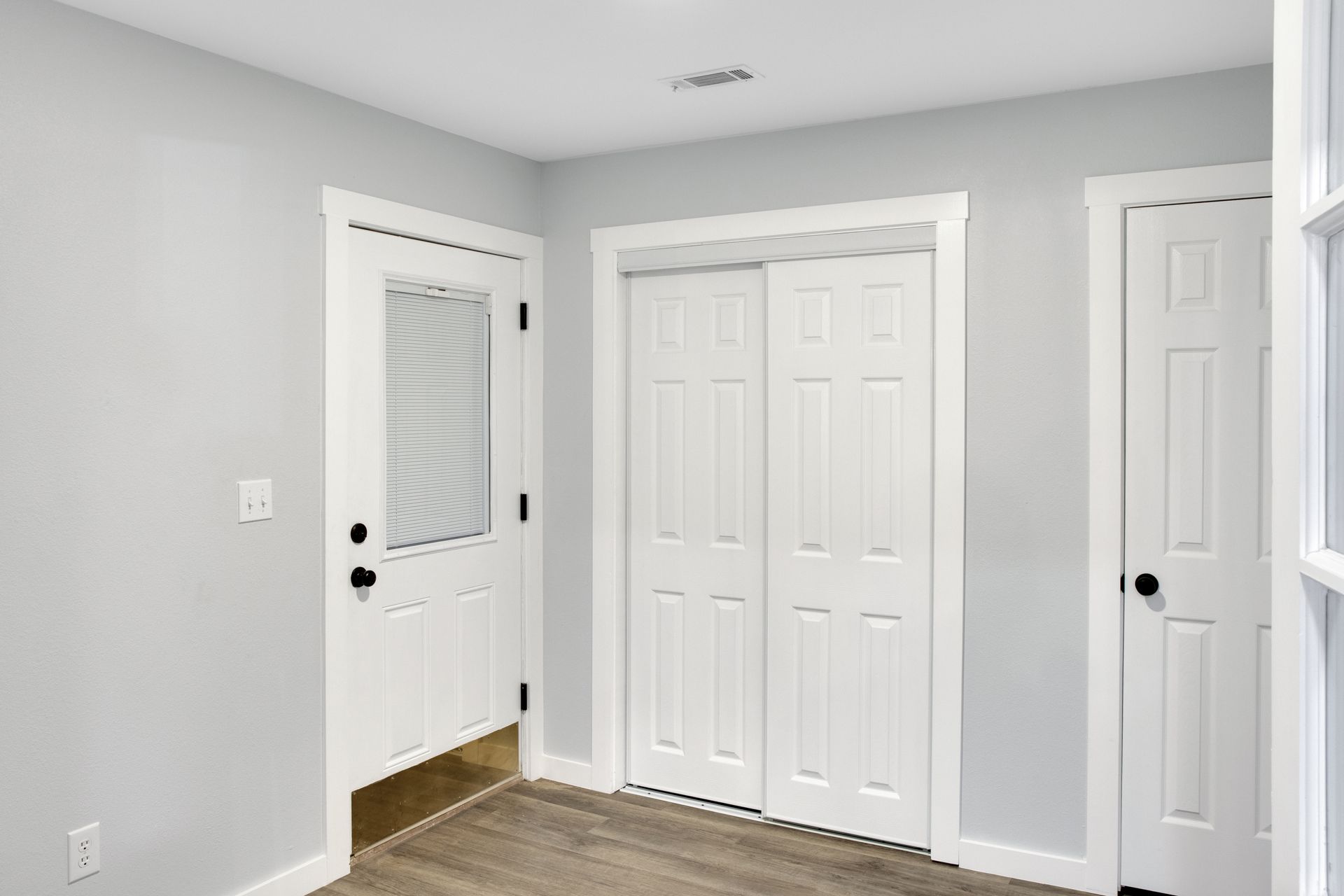 An interior view of a room with light gray walls, a white exterior door, a white double-door closet, and a white door.