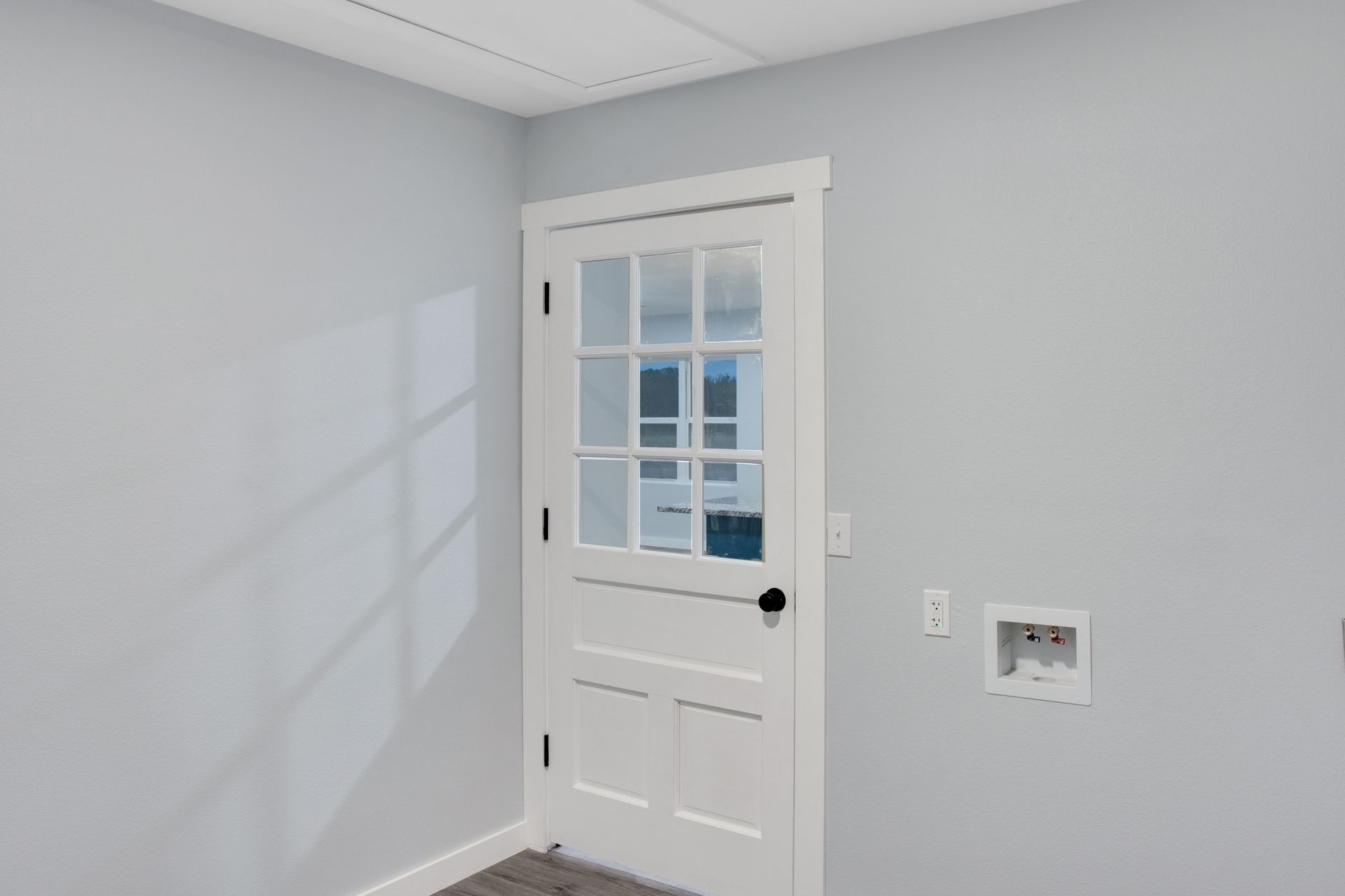 A white six-pane glass-panel door in a light gray room next to a recessed laundry utility box and a wall outlet.