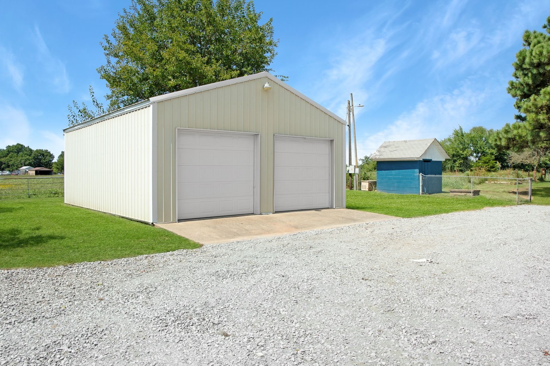 A beige metal two-car garage with white doors sits on a gravel driveway next to a small blue shed on a grassy lawn.