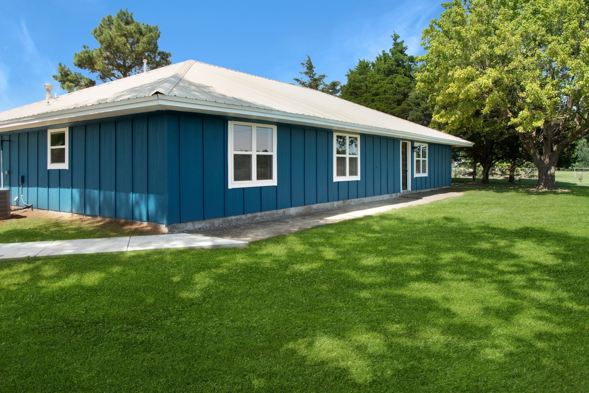 A one-story blue house with white trim and a white roof sits on a green lawn under a clear blue sky.