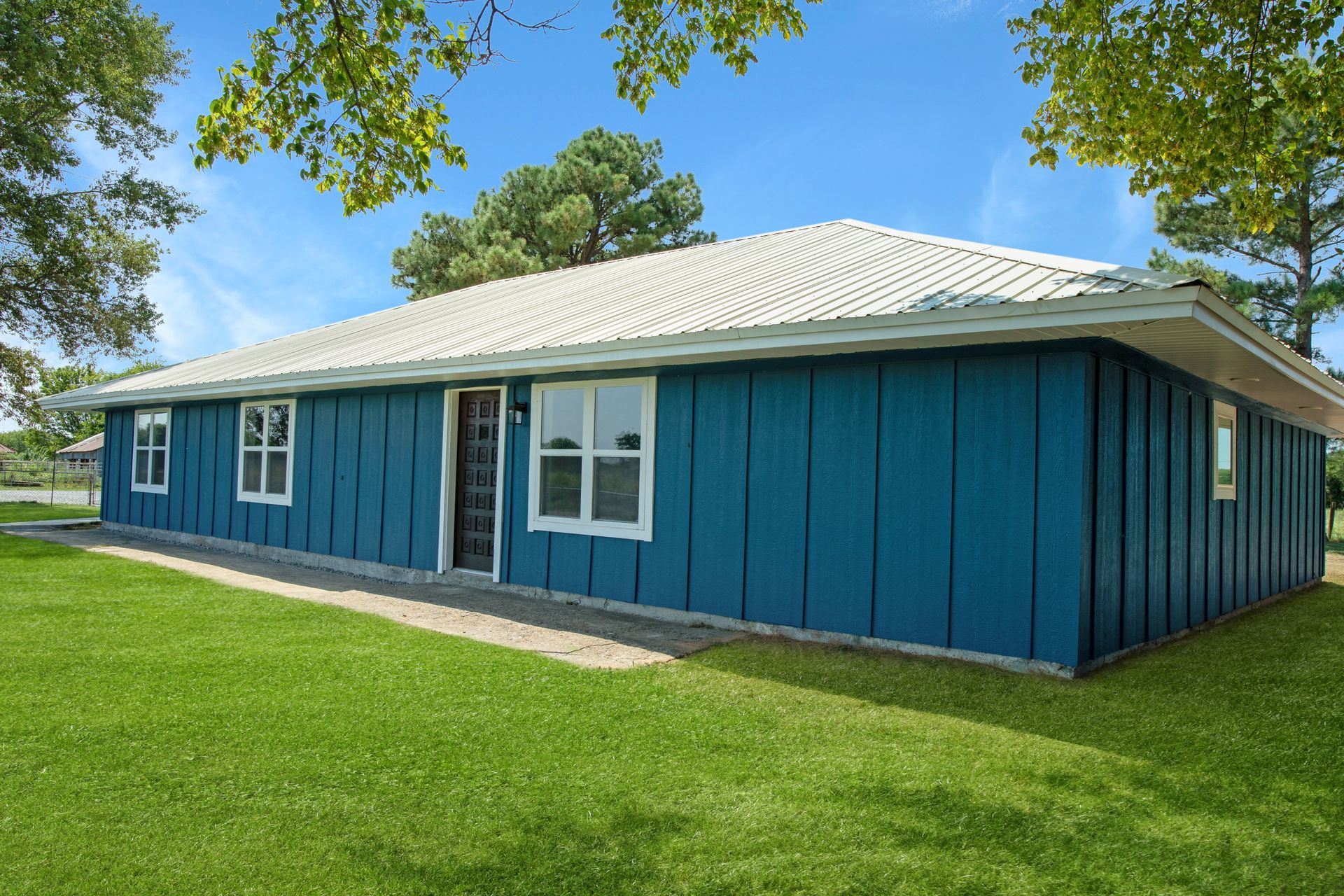 A single-story house with bright blue vertical siding and a white roof, situated on a green lawn under a sunny blue sky.
