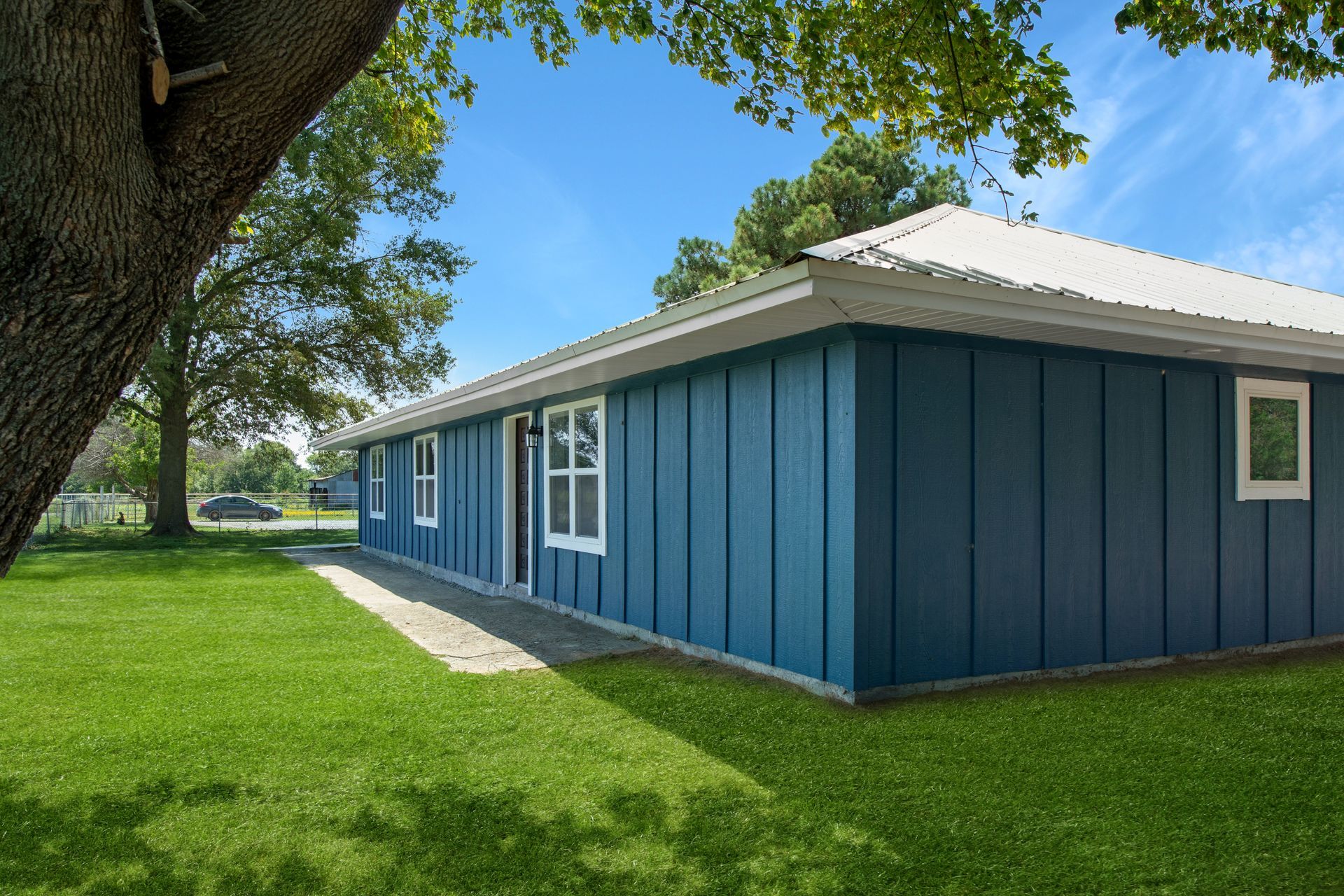 A blue, single-story house with vertical siding and a white roof sits on a green lawn next to a large tree.