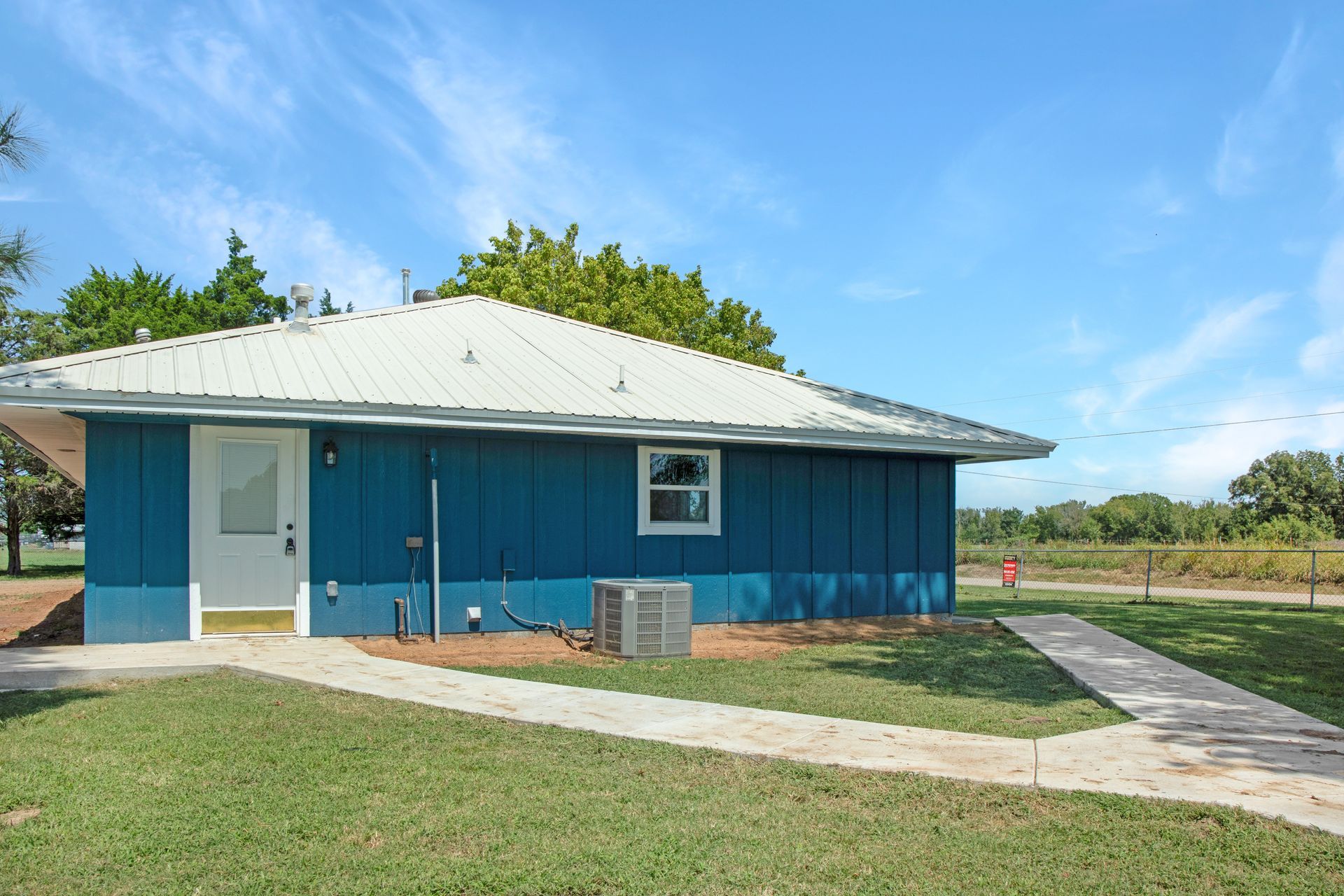 A blue one-story building with a white door and metal roof under a bright sky, surrounded by grass and concrete walkways.