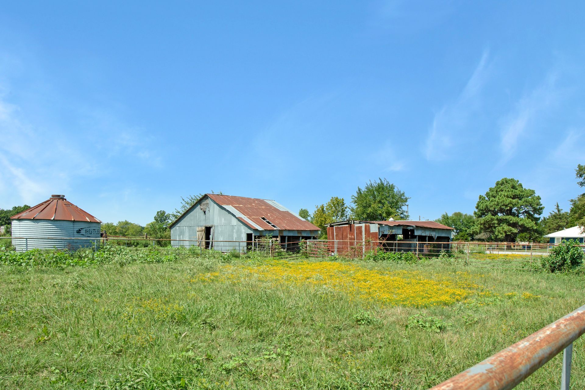 A rural farm scene with a metal grain silo, an aging barn, and smaller outbuildings set in a grassy field under a blue sky.