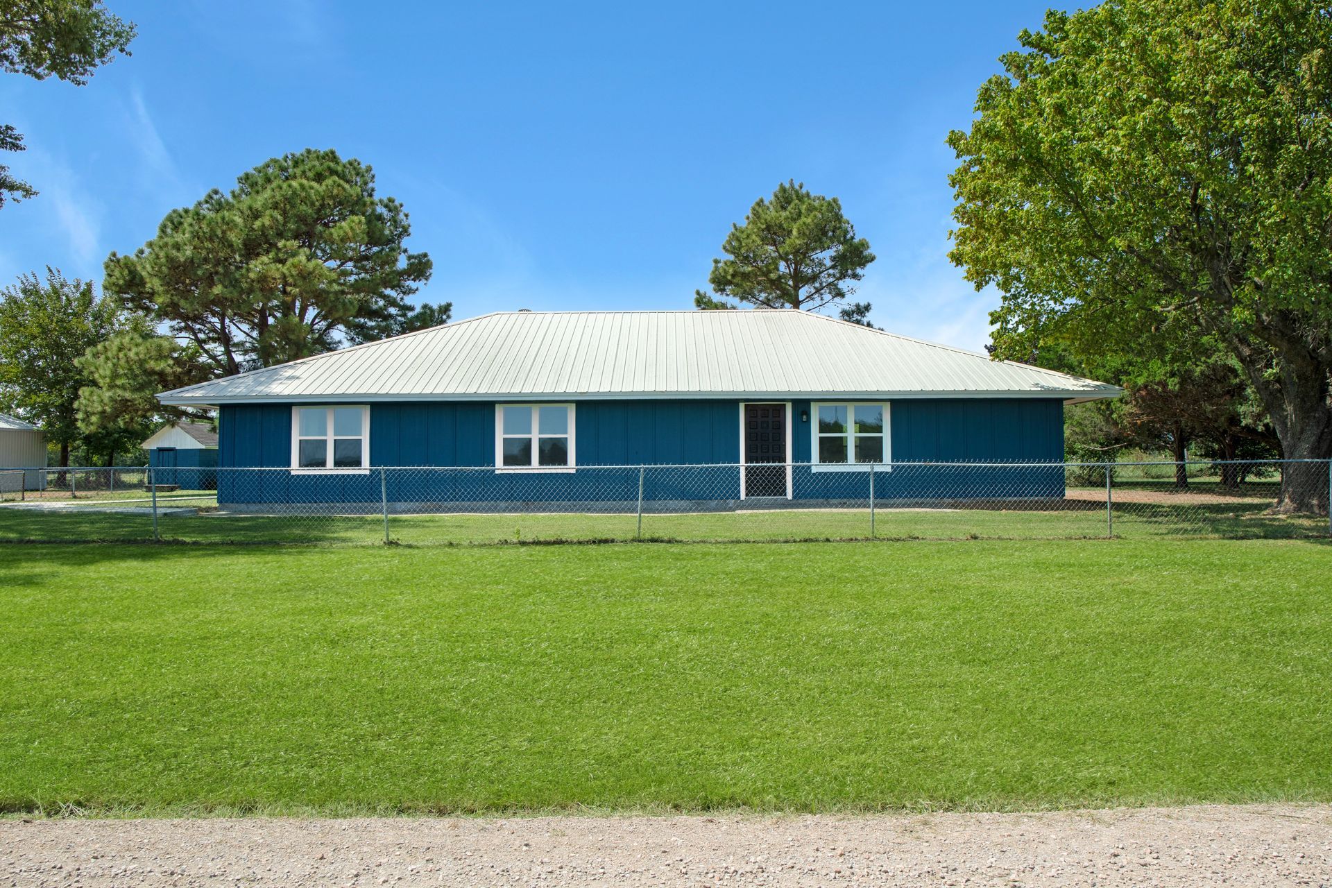 A single-story blue house with a white roof and metal fence, surrounded by green trees and a grassy lawn under a blue sky.