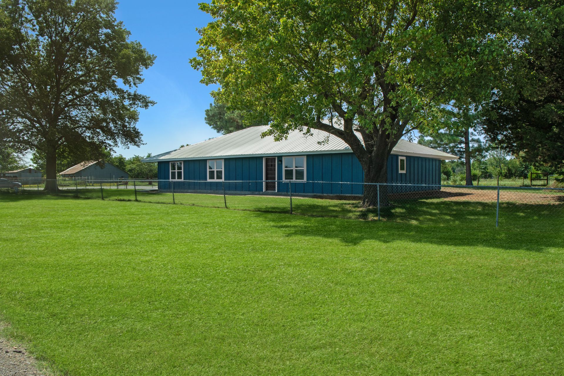 A blue, single-story house with a white metal roof stands behind a chain-link fence on a grassy, tree-filled lawn.