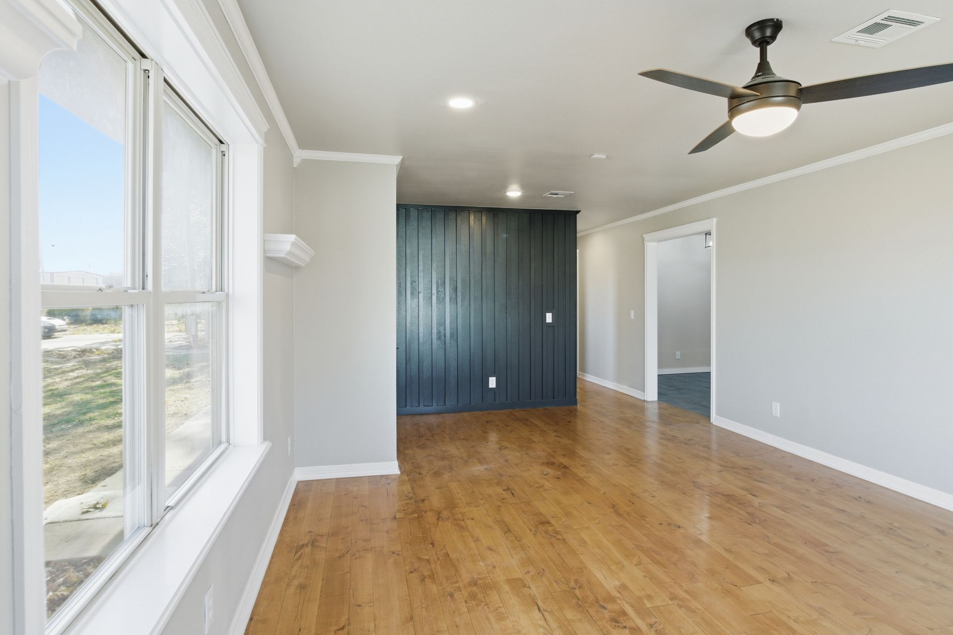 An empty room with light wood floors, gray walls, a large window, a dark wood accent wall, and a ceiling fan.