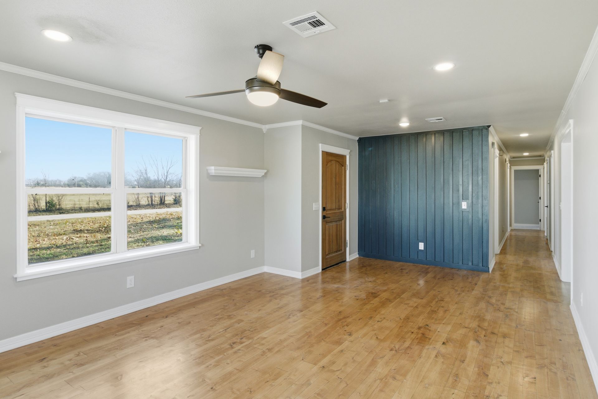 An empty room with light wood floors, gray walls, a large window, a ceiling fan, and a blue-paneled feature wall.