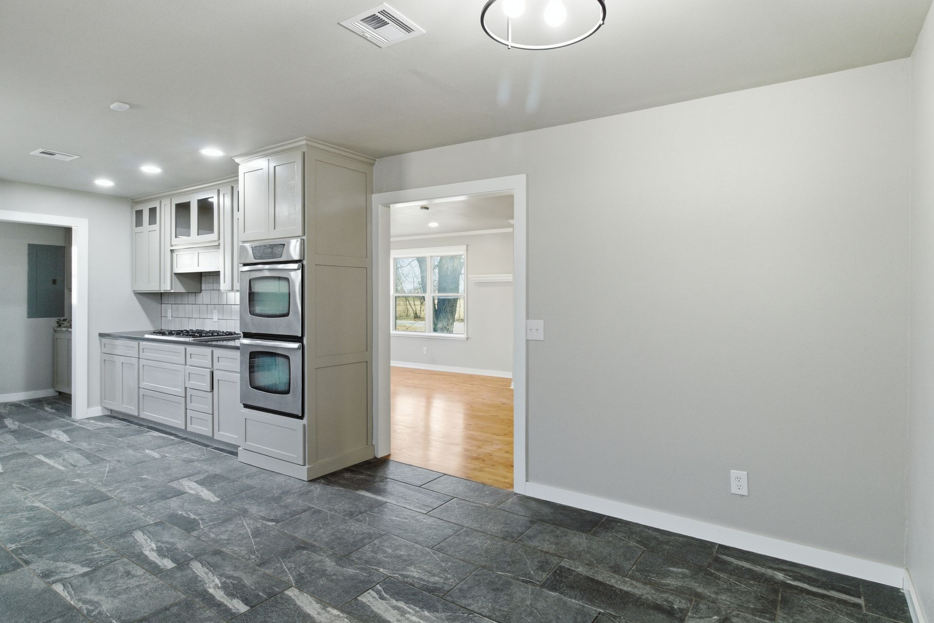 A modern kitchen interior featuring grey cabinets, built-in ovens, stone-look flooring, and a doorway to another room.