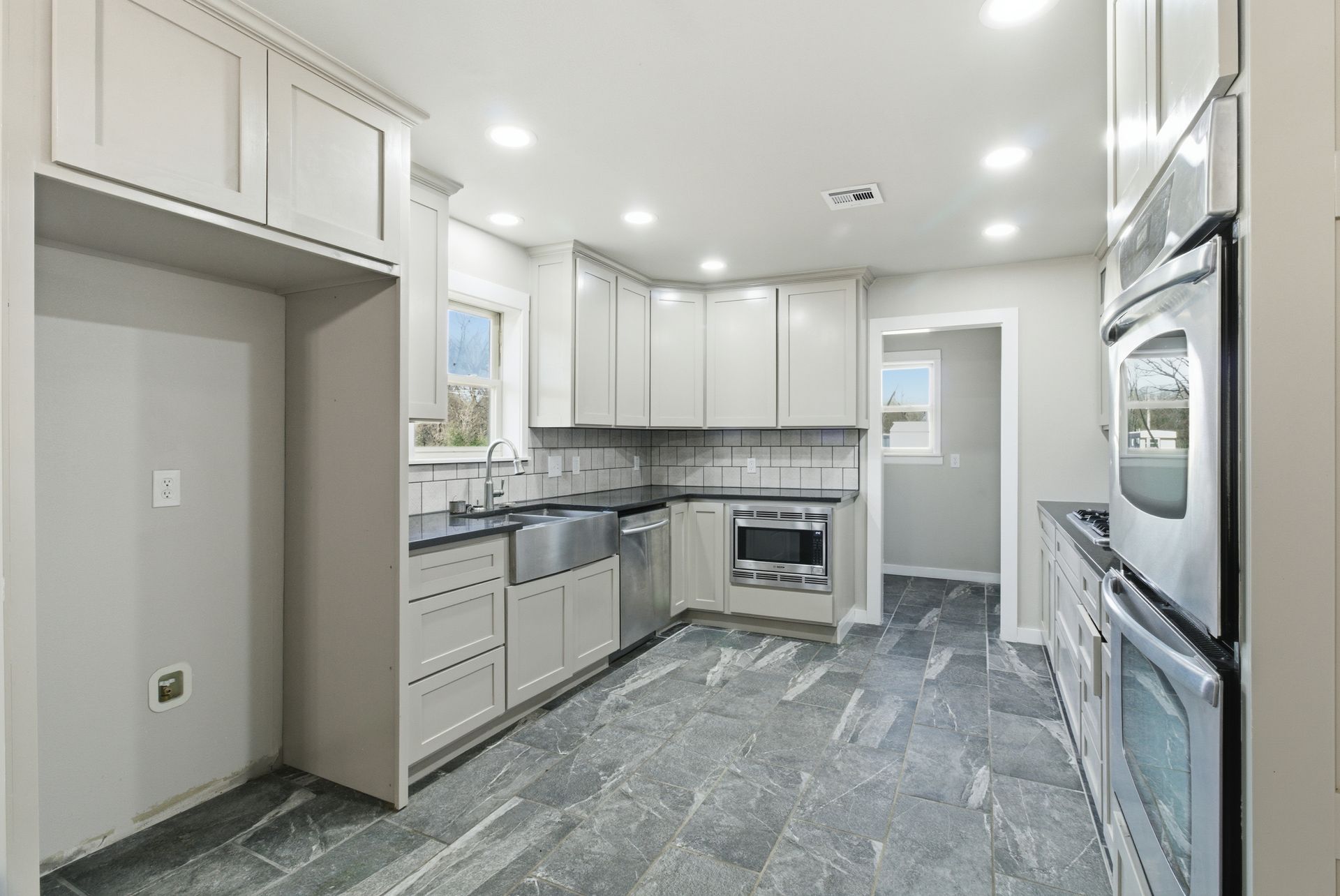 A modern kitchen with light gray cabinetry, stainless steel appliances, a farmhouse sink, and dark gray stone flooring.