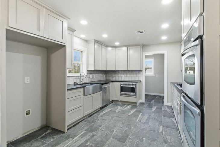 Modern kitchen with grey cabinets, dark stone flooring, stainless steel appliances, and a farmhouse sink.