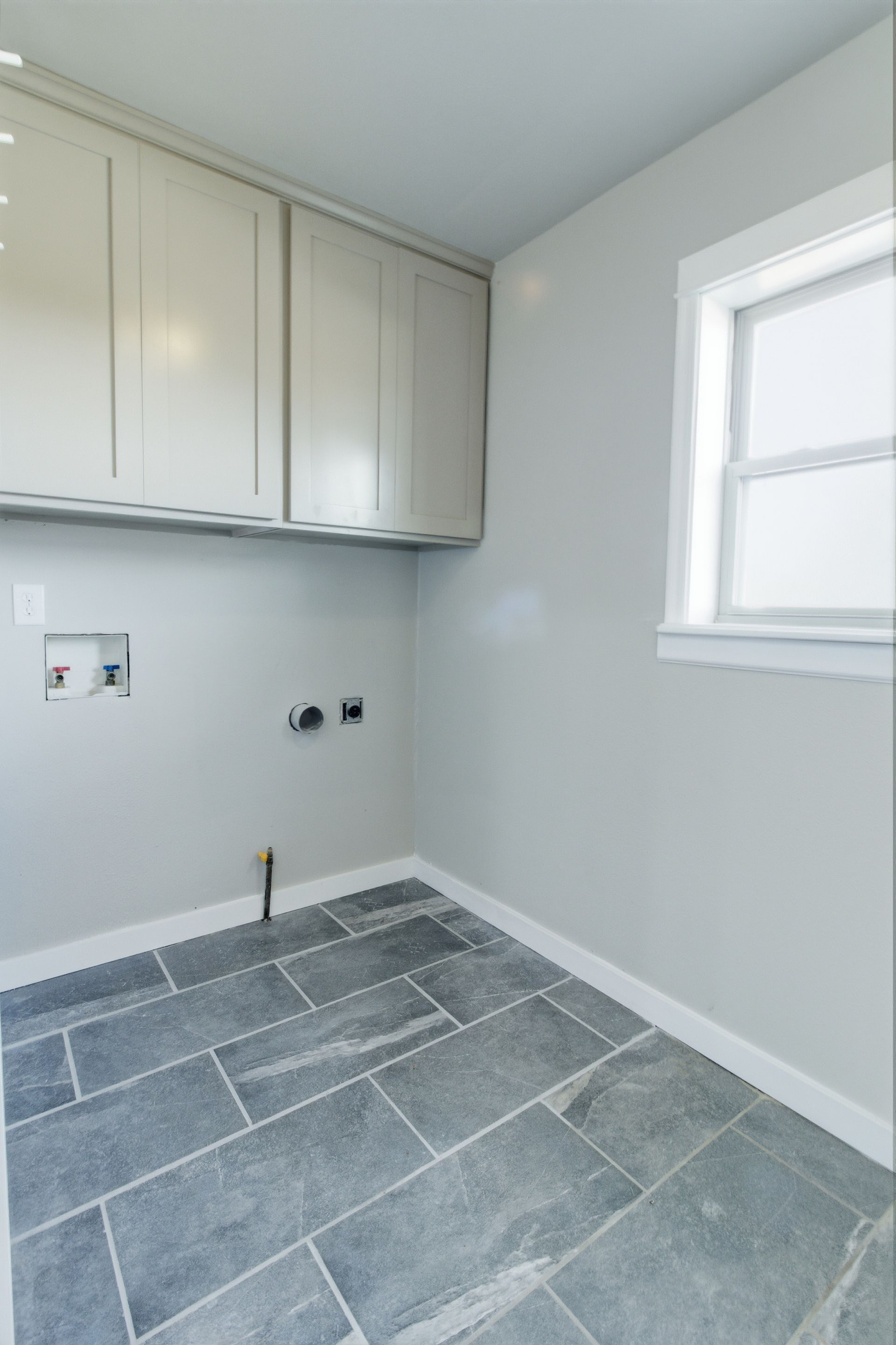 A laundry room with light gray walls, cream cabinets, and dark blue-gray stone flooring by a window.