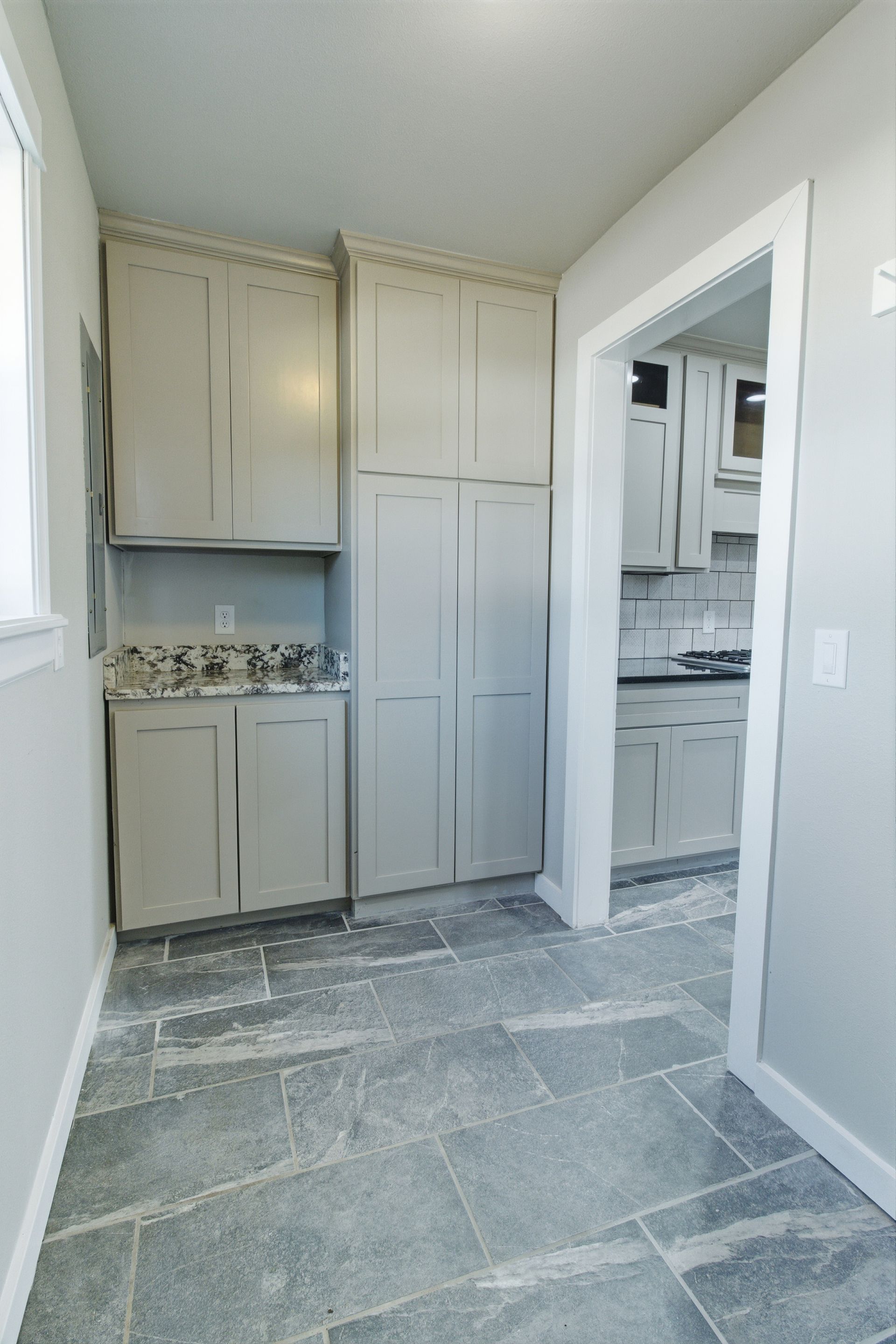 Gray built-in cabinets and a tall pantry unit in a hallway with gray stone tile flooring, leading to a kitchen.