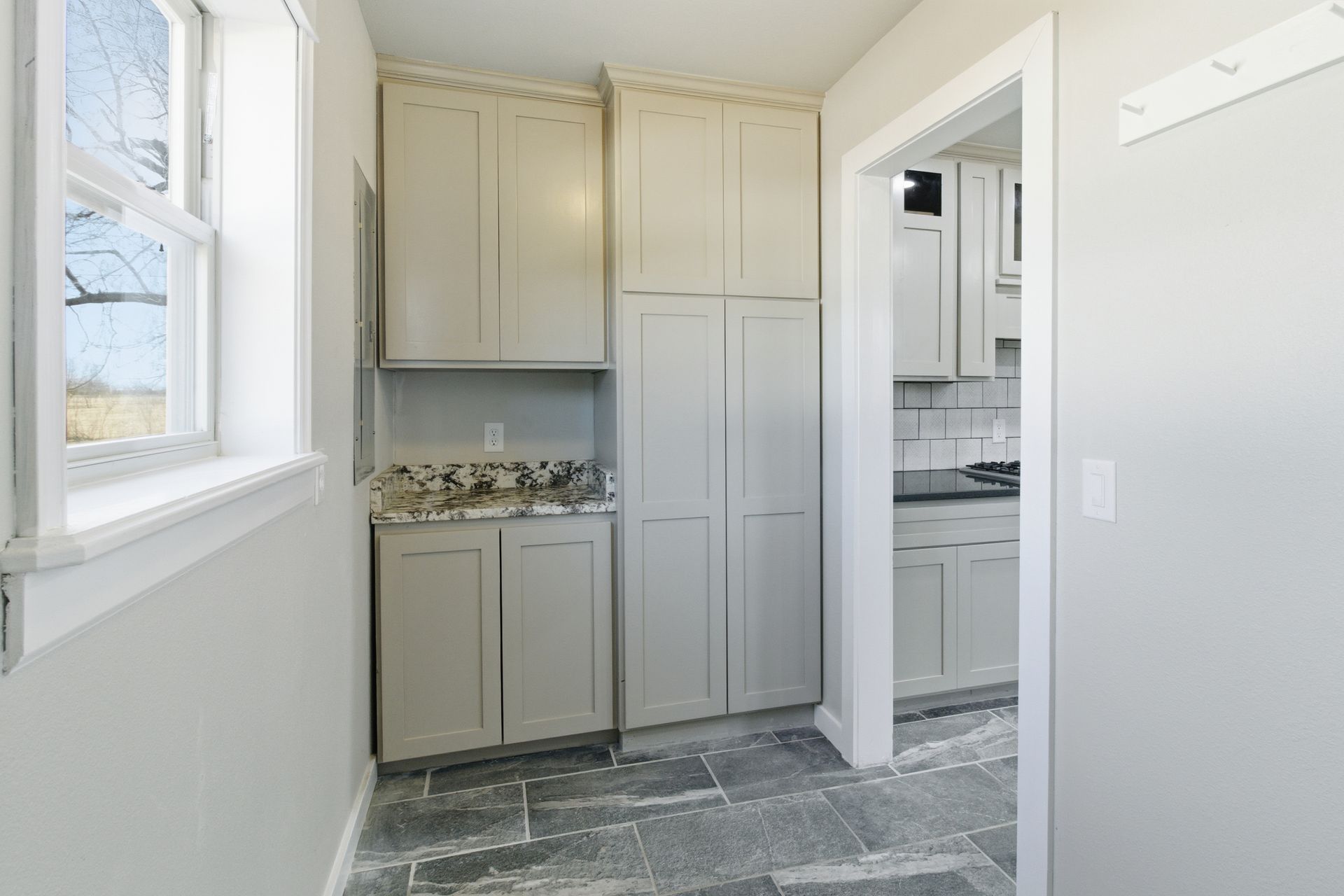 A mudroom with light gray cabinets, a granite countertop, and gray stone tile floors, leading into a kitchen.