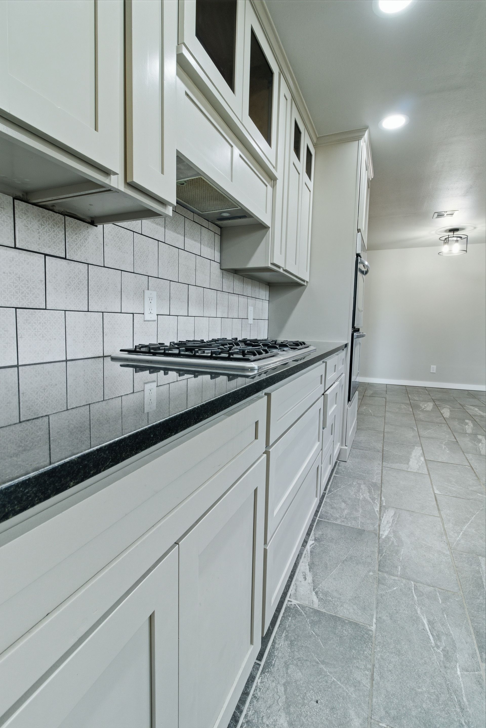 A bright kitchen featuring white cabinets, black countertops, a gas cooktop, gray tile backsplash, and stone flooring.