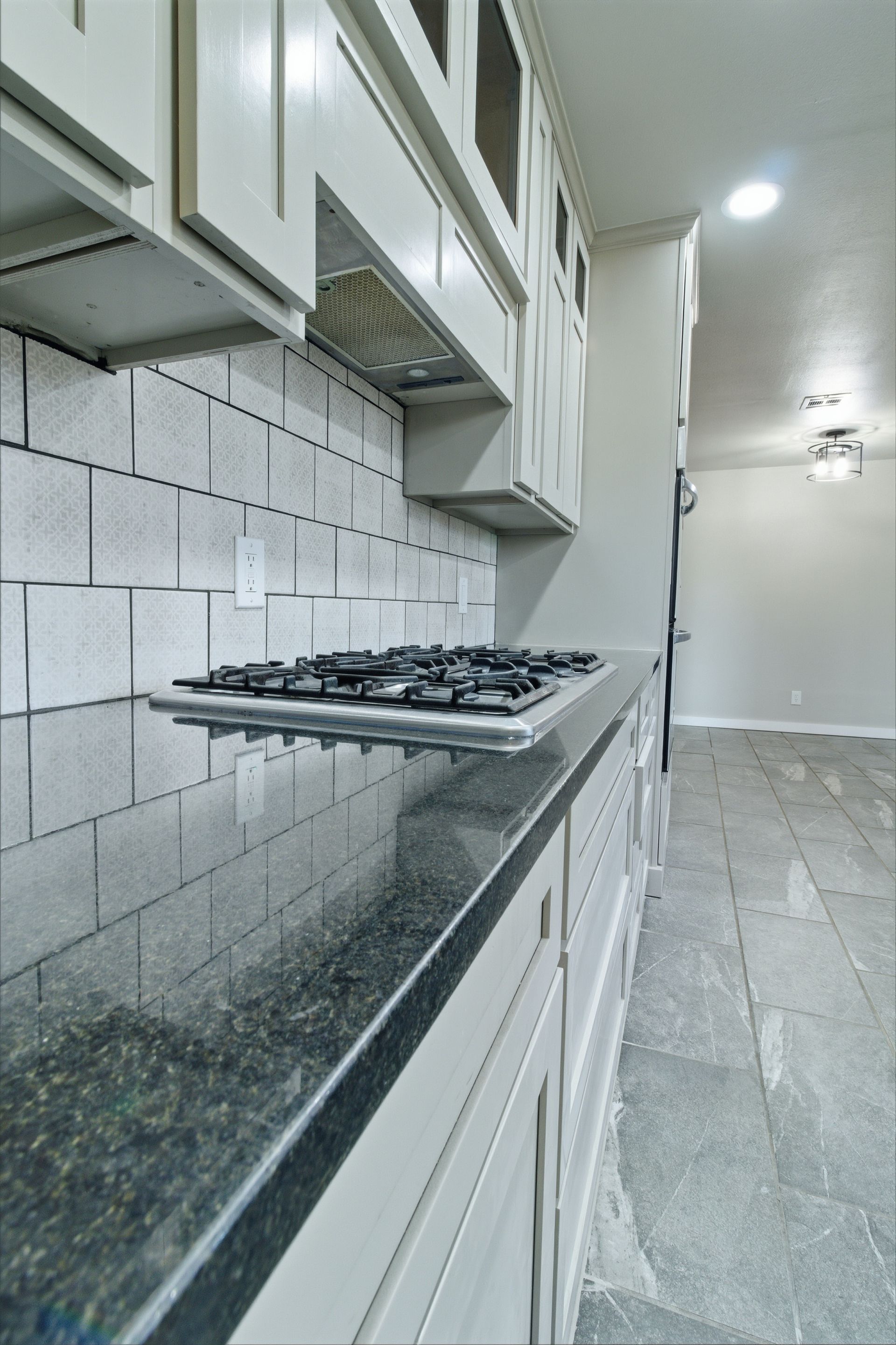 A modern kitchen featuring a gas stovetop built into dark granite countertops with light gray tile walls and cabinetry.