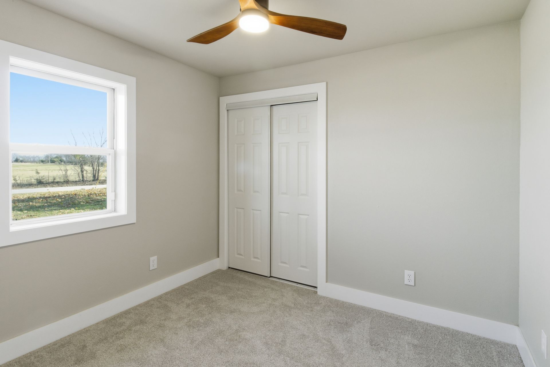 An empty room with light gray walls, carpeted flooring, a ceiling fan, a window, and a white two-door closet.